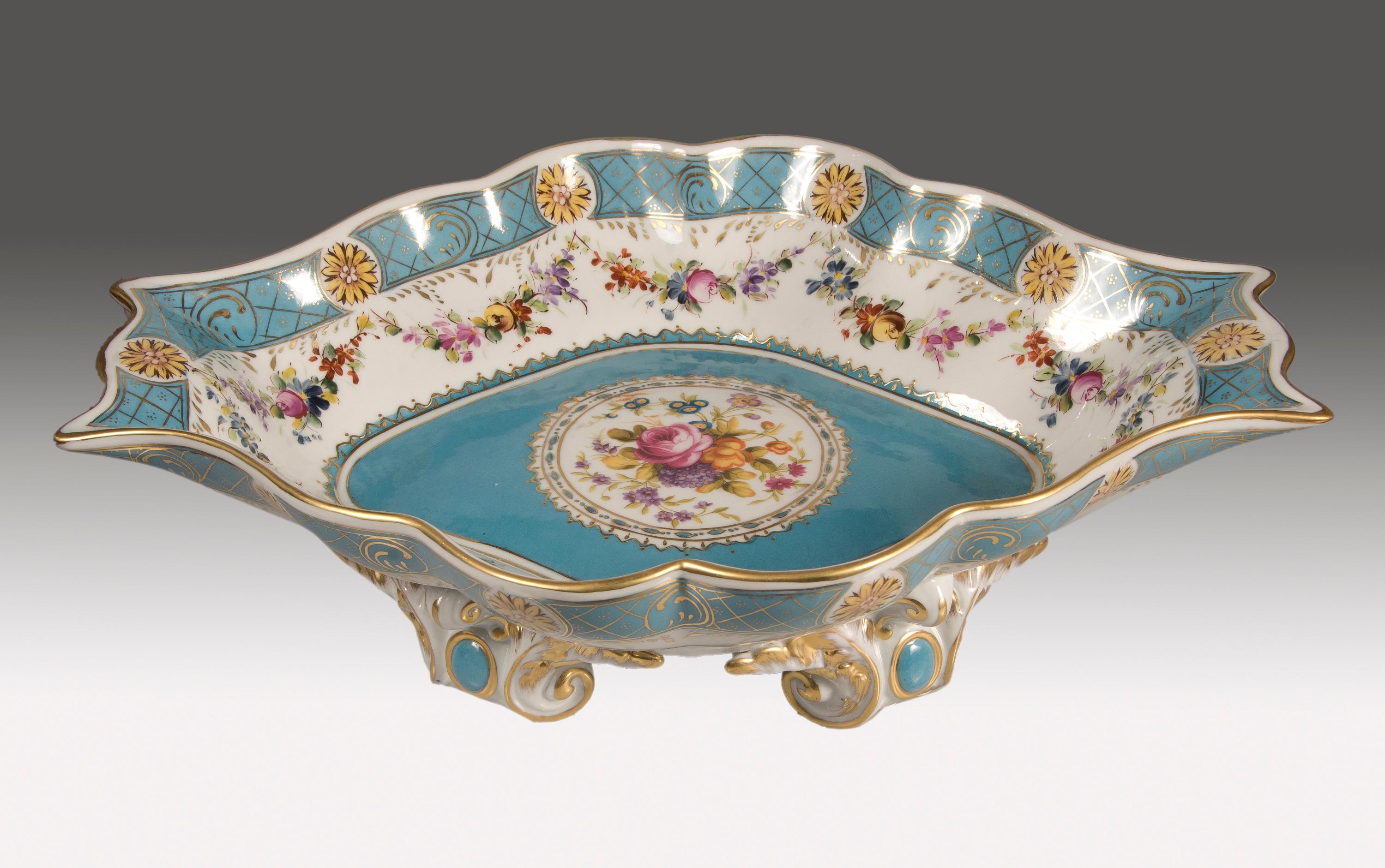 Centerpiece. Glazed porcelain.
Enameled porcelain centerpiece slightly raised on four volute-shaped legs and plain blue mirrors in front. The curved profiles, extended towards the neck, remind of examples inspired by the Rococo, while the
