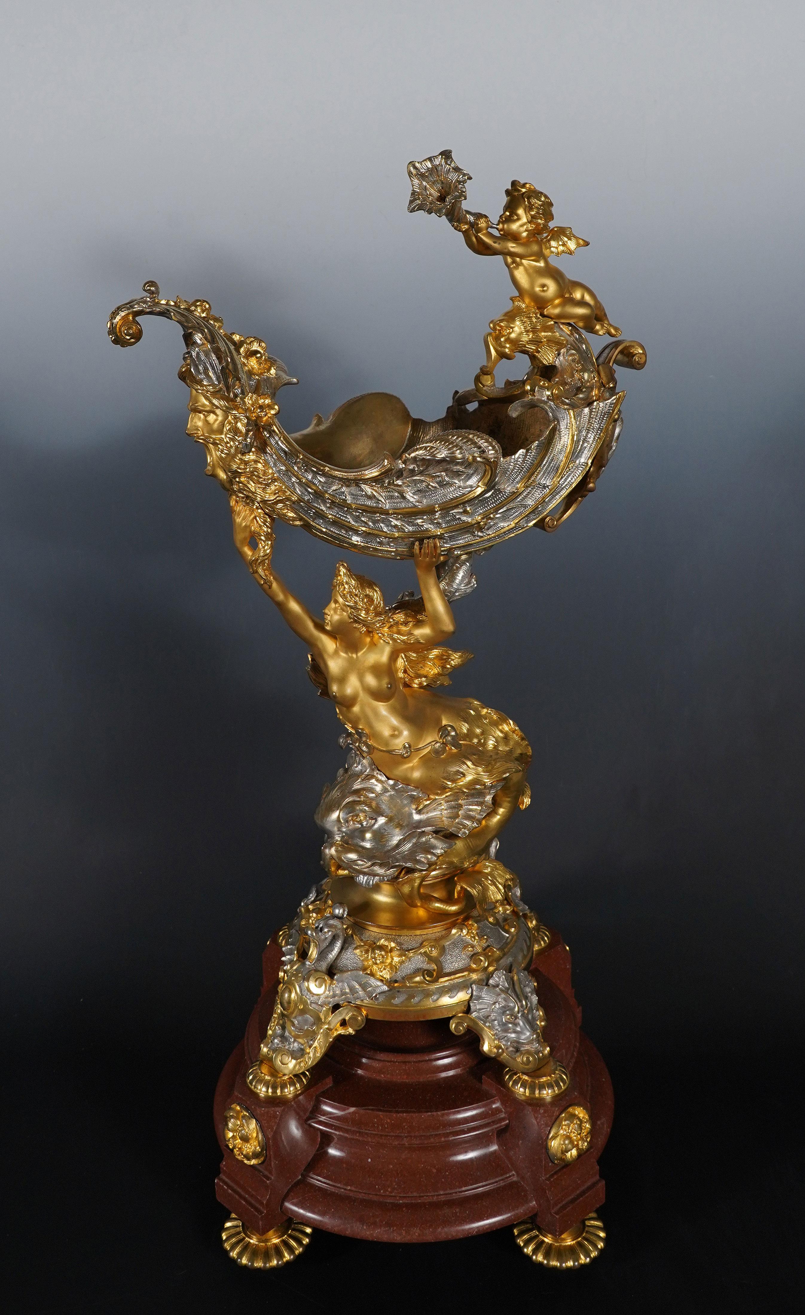 Exceptional centerpiece in silvered and gilded bronze, composed of a naiad riding a dolphin, supporting a chiseled shell decorated with reeds, adorned with the head of a river god on its bow and on which a winged love blows in a conch. The whole
