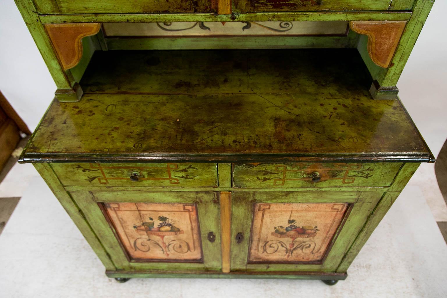 This cupboard has weathered green paint throughout with geometric and floral designs painted on the front. The lower doors have recessed panels with baskets of fruit supported by leaf arabesques. The upper doors have one fixed shelf as well as the