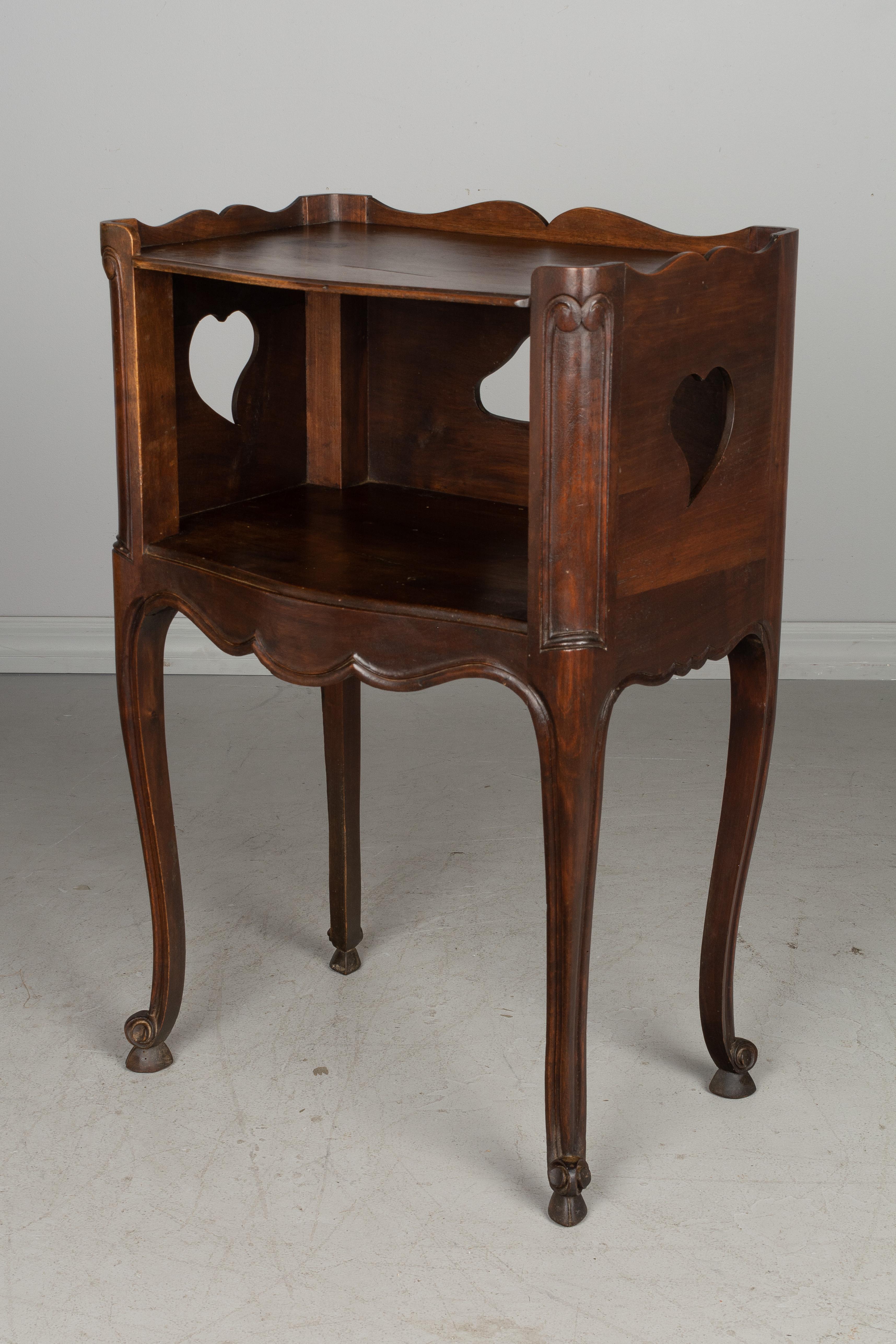 A Louis XV style Country French side table or nightstand made of solid walnut, with open niche and pierced cut-out shapes on the back and sides. Scalloped apron and gallery surrounding the top. Curved corners and slender legs with scroll feet. Waxed