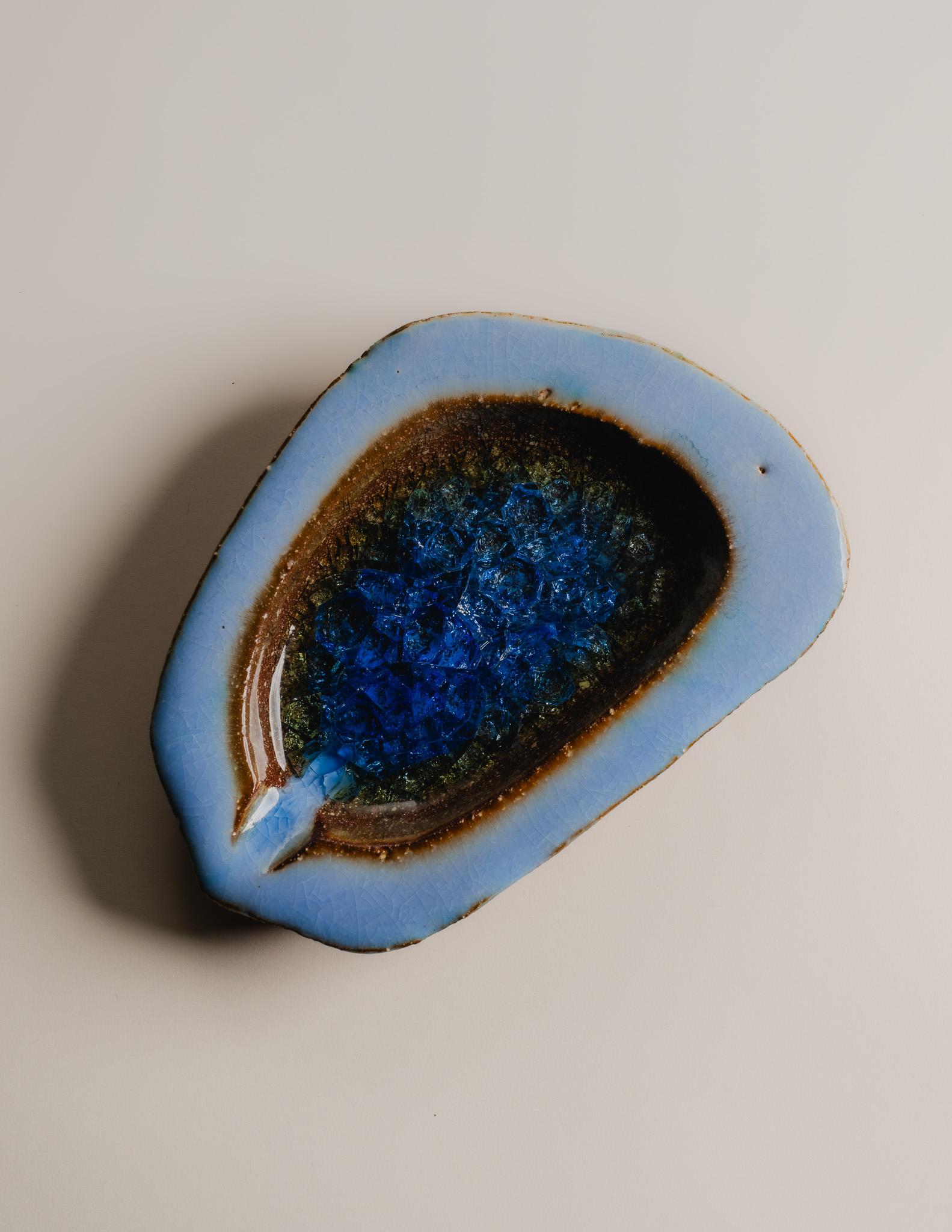 Aschenbecher aus blau glasierter Keramik mit verschmolzenem Glas in der Mitte, 1970er Jahre.
Mit einem Einschluss aus geschmolzenem Glas in tiefblauen und bernsteinfarbenen Tönen, der einen knisternden, durchscheinenden Effekt erzeugt, der an eine