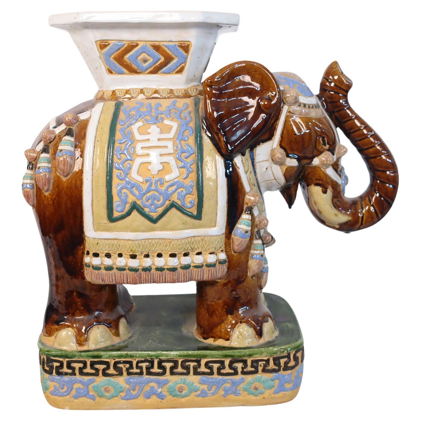 Single ceramic brown Asian elephant garden stool with brown yellow and white color and gold, green, and light lilac saddles and trim. The stool portrays an elephant with raised trunk. This adorable stool is perfect to set your drink down on as a