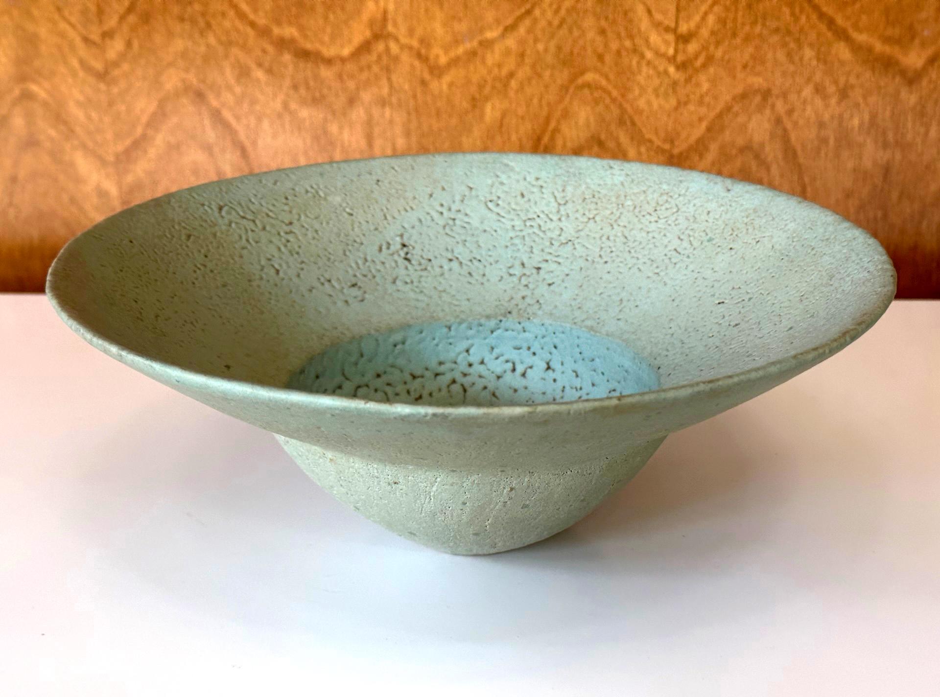 A stoneware glazed bowl by British studio potter John Ward (1938-2023) circa 1980-1990s of the last quarter of the 20th century. The bowl features a distinct form with a wide flanged rim and spherical body. The surface is covered with a copper green