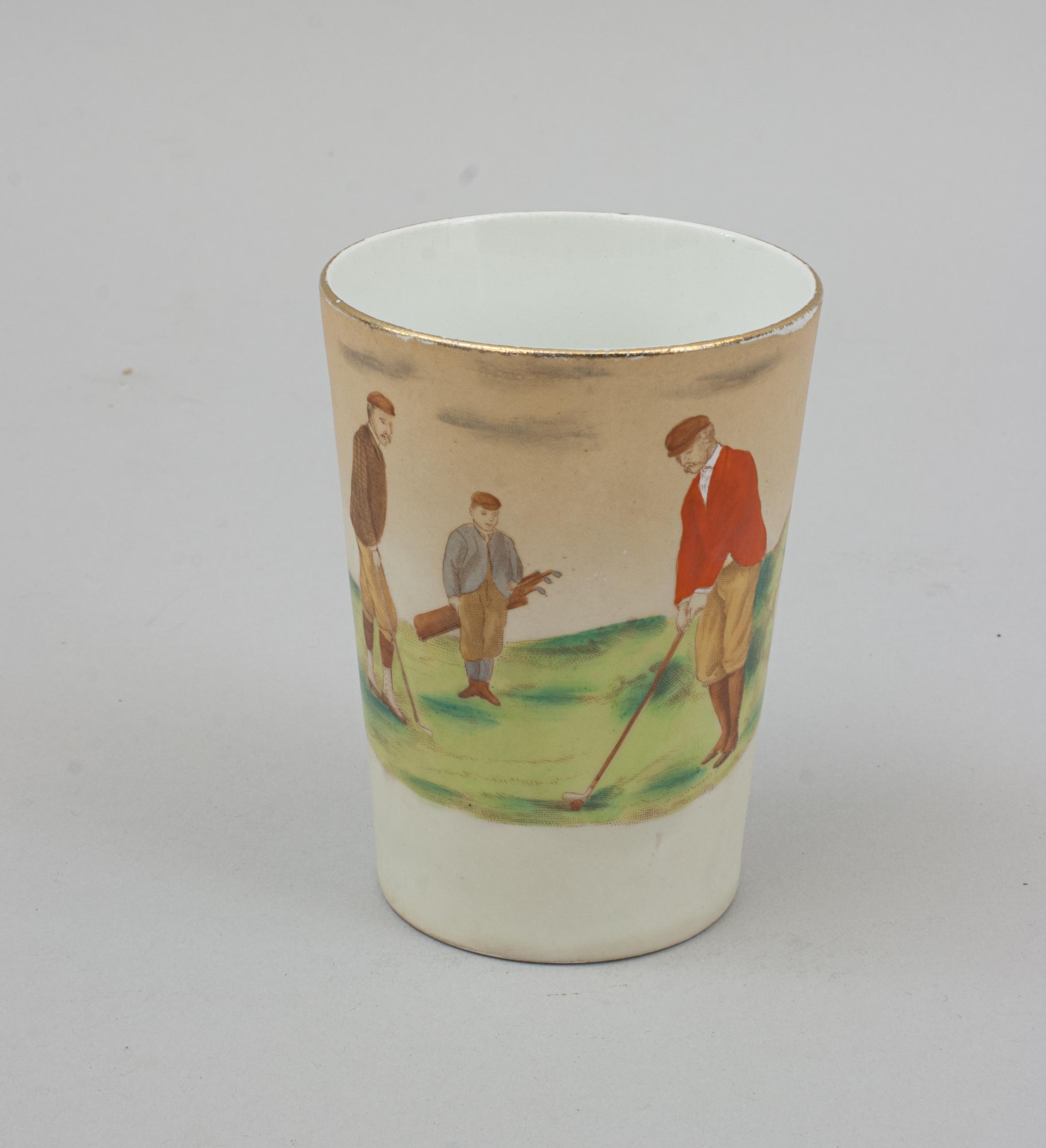 Wiltshaw & Robinson Carlton Ware Golf Beaker.
A ceramic Carlton Ware beaker with a golfing scene and gilt rim. The golf scene shows one golfer dressed in a red coat about to strike the ball watched by another player and a caddy. The bottom on the