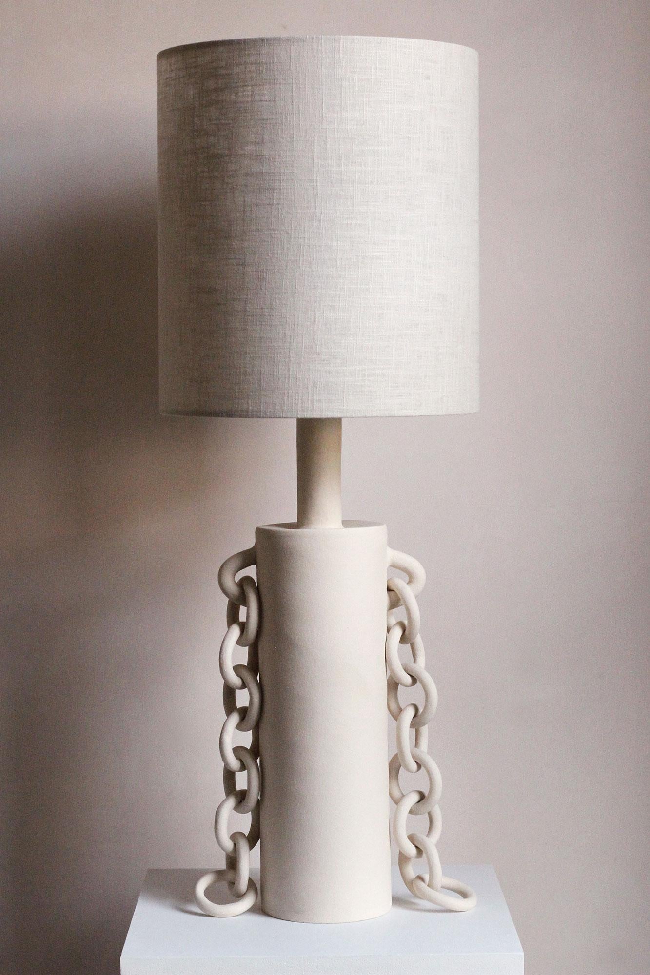 Handmade from stoneware, this contemporary table lamp features two ceramic chains that hang either side of the cylindrical lamp base, adorning the form like jewellery. The tall cream/grey linen shade is handmade in the UK, the three cylindrical