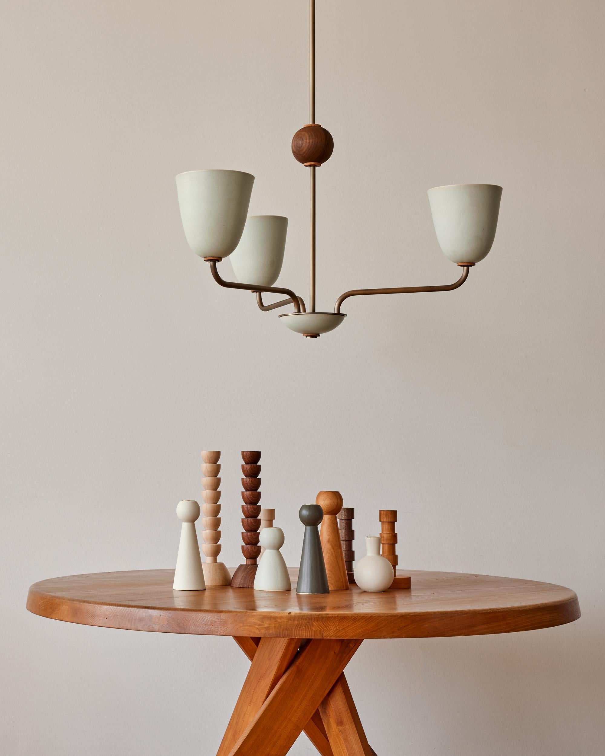 Three antique brass patina arms curve up from a ceramic cluster dish to three tall stoneware ceramic shades in a matte glaze with a tinge of blue. With a fixed black walnut orb and tan harness leather accents, this graceful chandelier has the