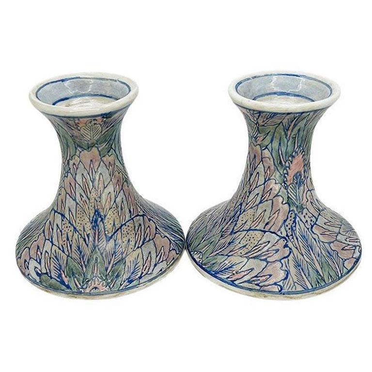 A pair of ceramic pastel pink, blue and purple candlestick holders. Each piece is round and created from ceramic. The exterior of each candle holder is decorated in a flame stitch look design in muted pinks, purples, and blues. Each is stamped at