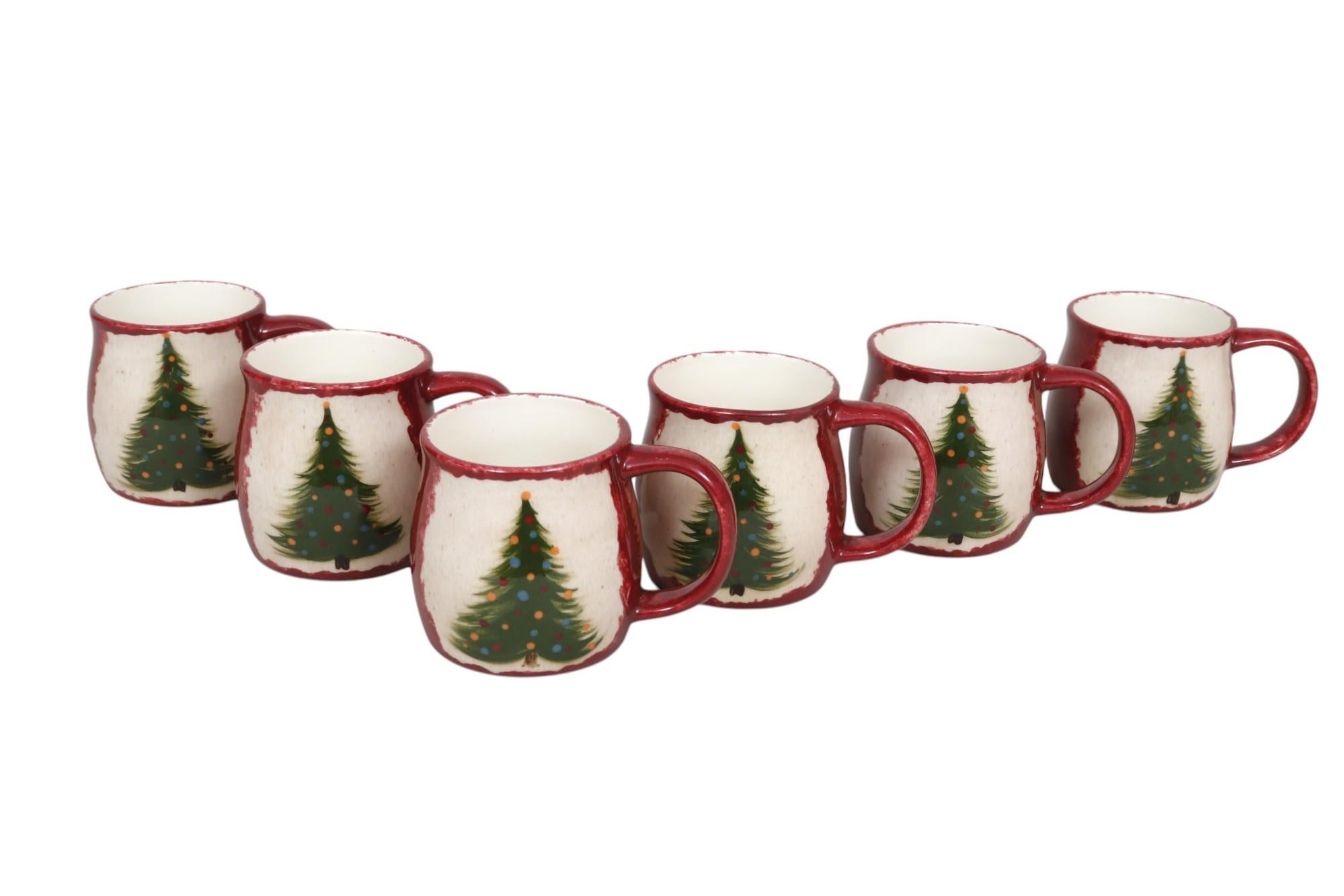 A set of six ceramic festive Christmas mugs. Each is individually hand-painted with a brushed green Christmas tree. Red, blue, and gold baubles decorate the trees, set against a cream background. The body of the mugs and the handles are sponged red.