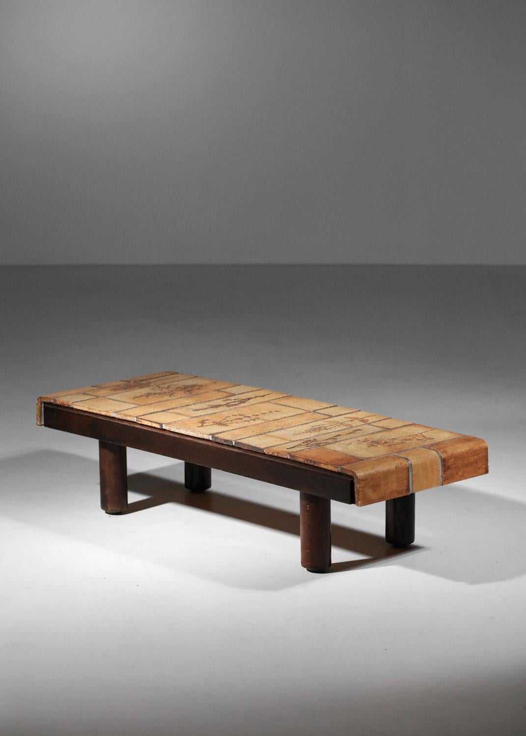 Table basse française des années 50 par l'artiste Roger Capron. Base en bois massif et plateau composé de carreaux de céramique de différentes tailles émaillés dans des teintes beiges et brunes. La table présente le motif d'herbier typique de