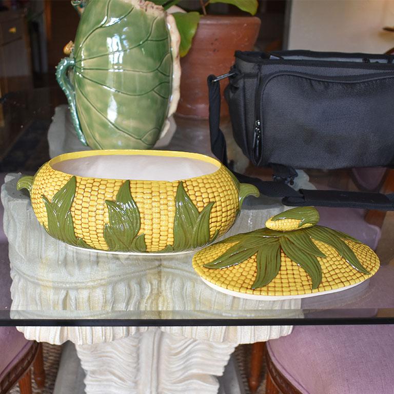 A hand-painted ceramic soup tureen is shaped like a bushel of yellow corn with green leaves. This oval serving dish is created from ceramic, and hand-painted by the artist. The sides and top are textured to resemble corn kernels. The lid at the top