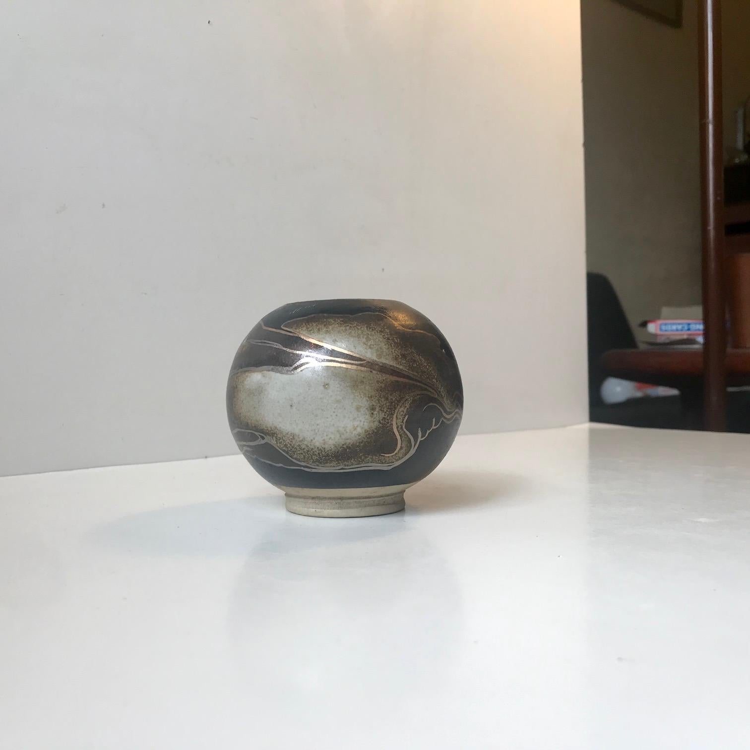Peter Sylvest has worked at both Royal Copenhagen and Michael Andersen in Denmark and has exhibited all-over the world. This small footed ball vase has a flowing abstract decor in earthy glaze sand is highlighted with gold and silver only. The vase