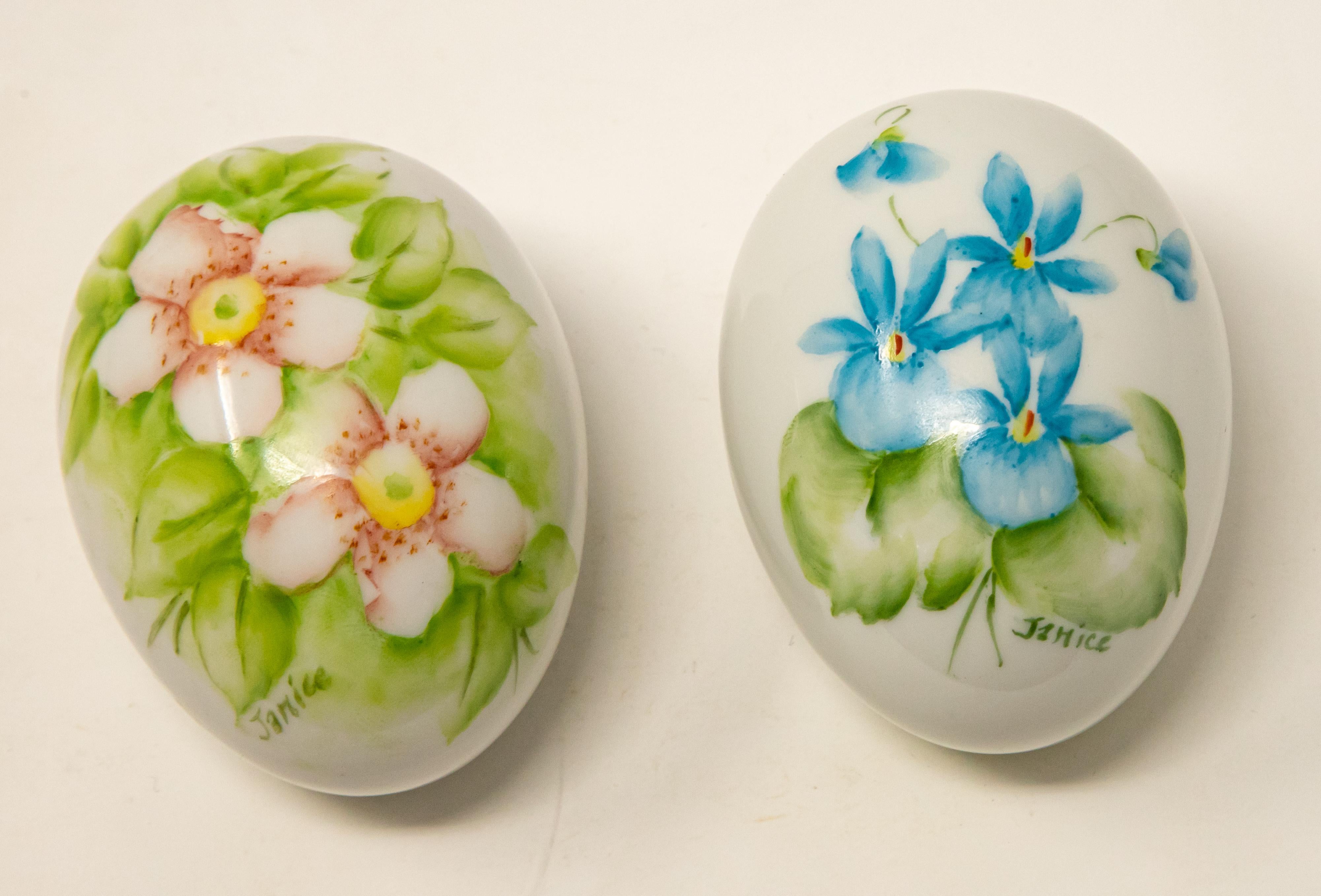 Offering a pair of ceramic hand painted eggs. Both are cream in color. One has light pink florals with greenery around them, and the other is blue florals with greenery. Both are signed Janice, and the bottoms are marked, hand painted by Janice