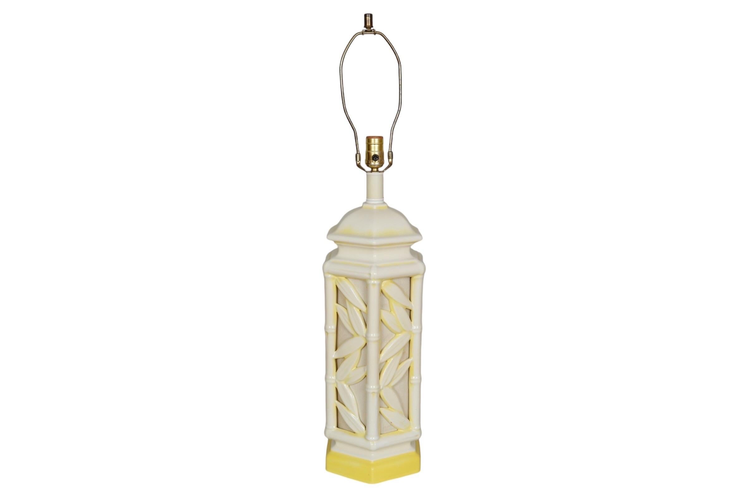 A ceramic faux bamboo table lamp in off white with a yellow base. The hexagonal body is shaped like a lantern made of bamboo, with bamboo leaves adorning the linen covered sides. Measures 25