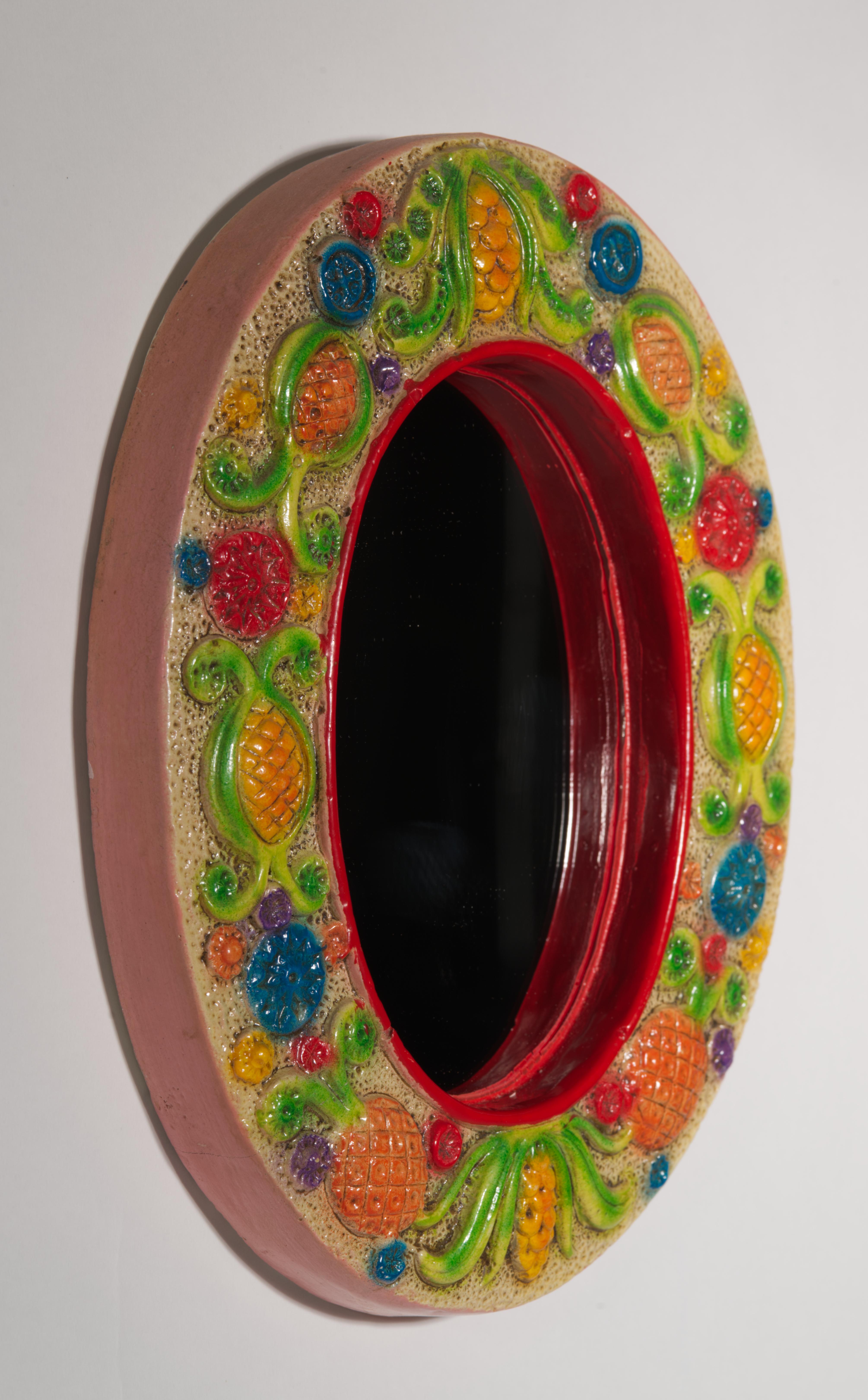  Il piccolo specchio da parete vintage è caratterizzato da una cornice in ceramica colorata e giocosa, decorata con una fascia di ananas, fiori e foglie nei colori arancio, verde, rosso e blu su sfondo giallo. Il bordo interno è decorato con un