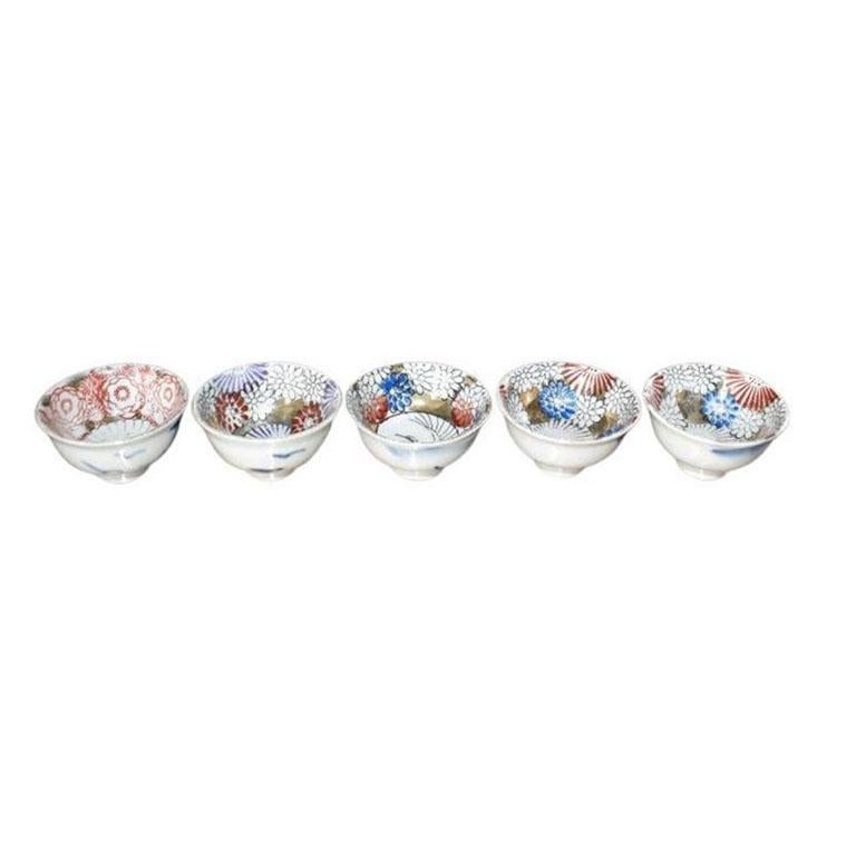 A set of five ceramic handpainted Japanese sake cups. Each cup is round and sits upon a delicate footed base. Each has been hand-painted on the inside with blue, white, and deep red chrysanthemums, and is accented in gold. The center of one of the