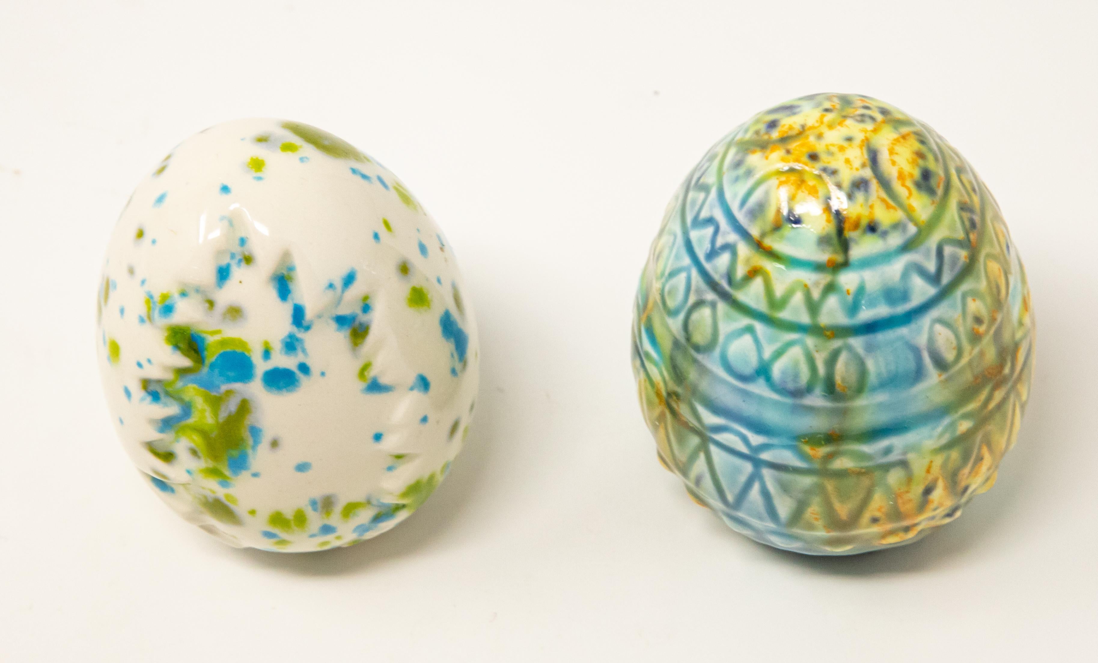 Offering a pair of ceramic glazed eggs. One egg is ivory, with blue and green spots with a tree stamped into it. The other is mostly blues with accents of green and yellow, and has geometric designs around the sides.