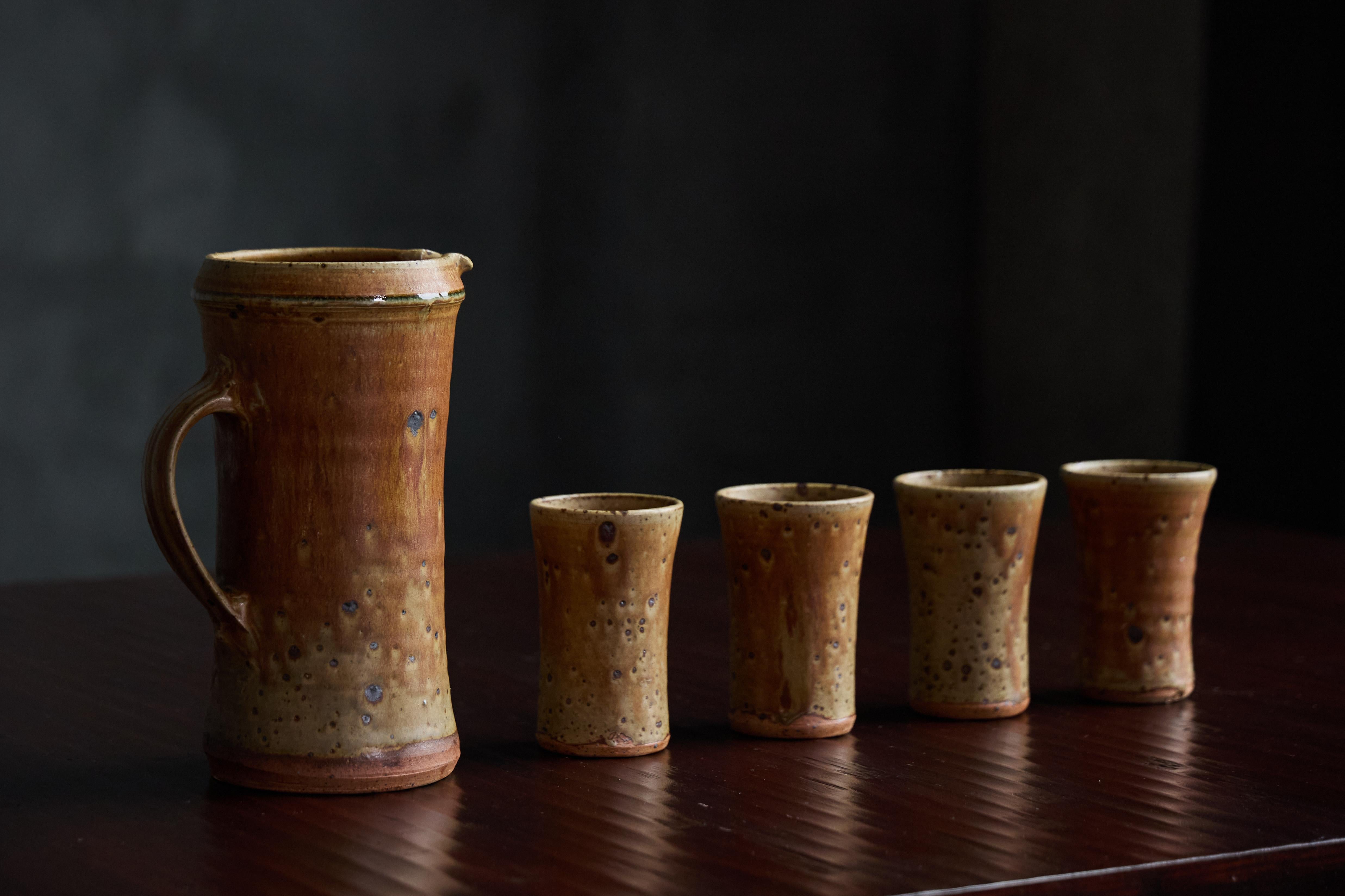Elegant stoneware set by French ceramic artist Gustave Tiffoche featuring a large, earthy-toned pitcher paired with four uniquely shaped goblets, exuding a rustic charm characteristic of mid-20th century design. Each piece showcases Tiffoche’s