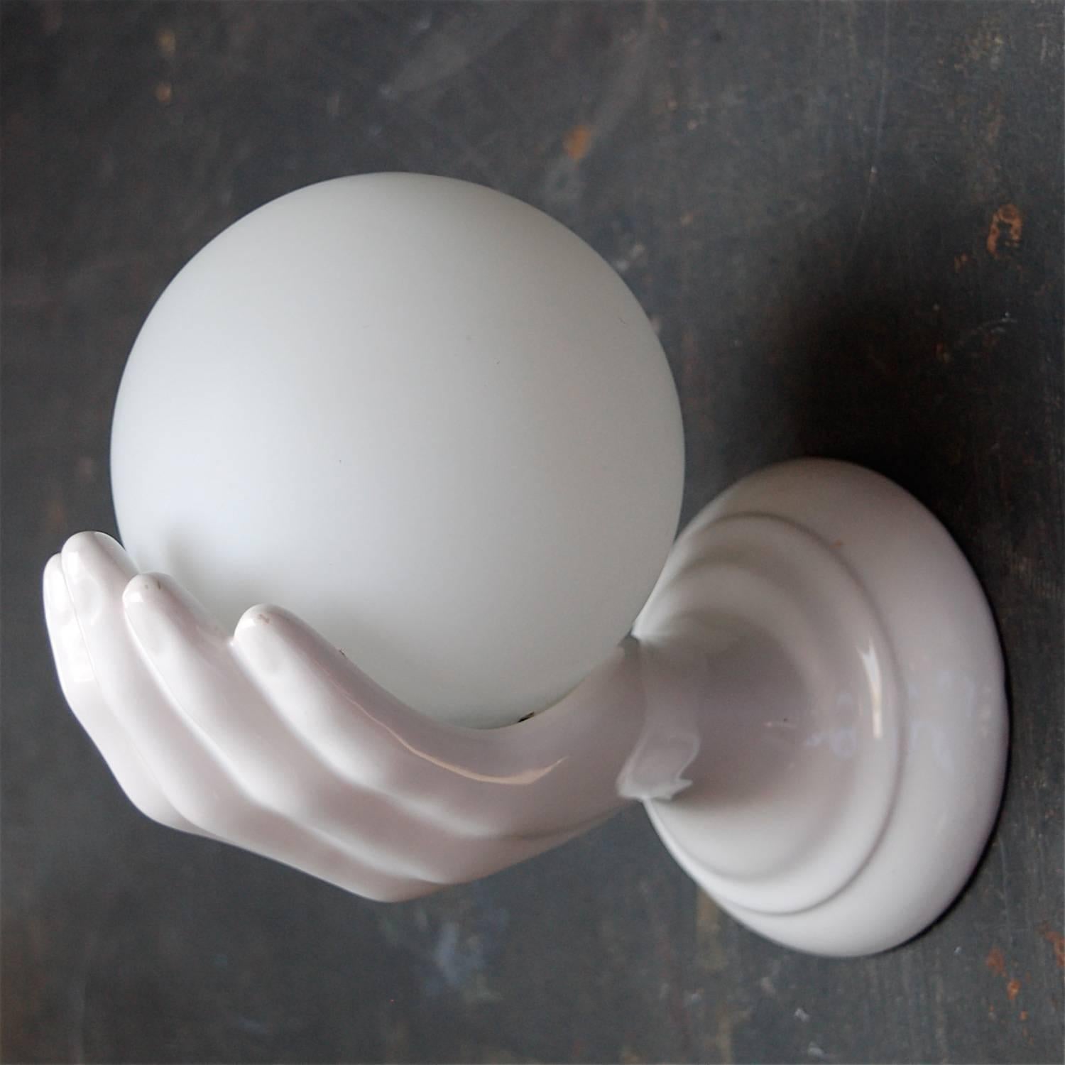 Vintage 1980s Italian ceramic wall lamp in form of a glossy white hand, cupping or holding a white glass globe with matt finish. The globe is held in place by clasps hidden from view. There are no chips or cracks to either the porcelain or glass.