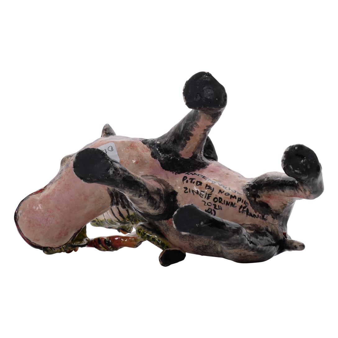 Esta juguetona escultura de cerámica captura un caprichoso momento del corazón de la naturaleza africana: una jubilosa rana encaramada triunfalmente sobre el lomo de un hipopótamo, con una hoja en la mano como si encabezara un desfile. Totalmente