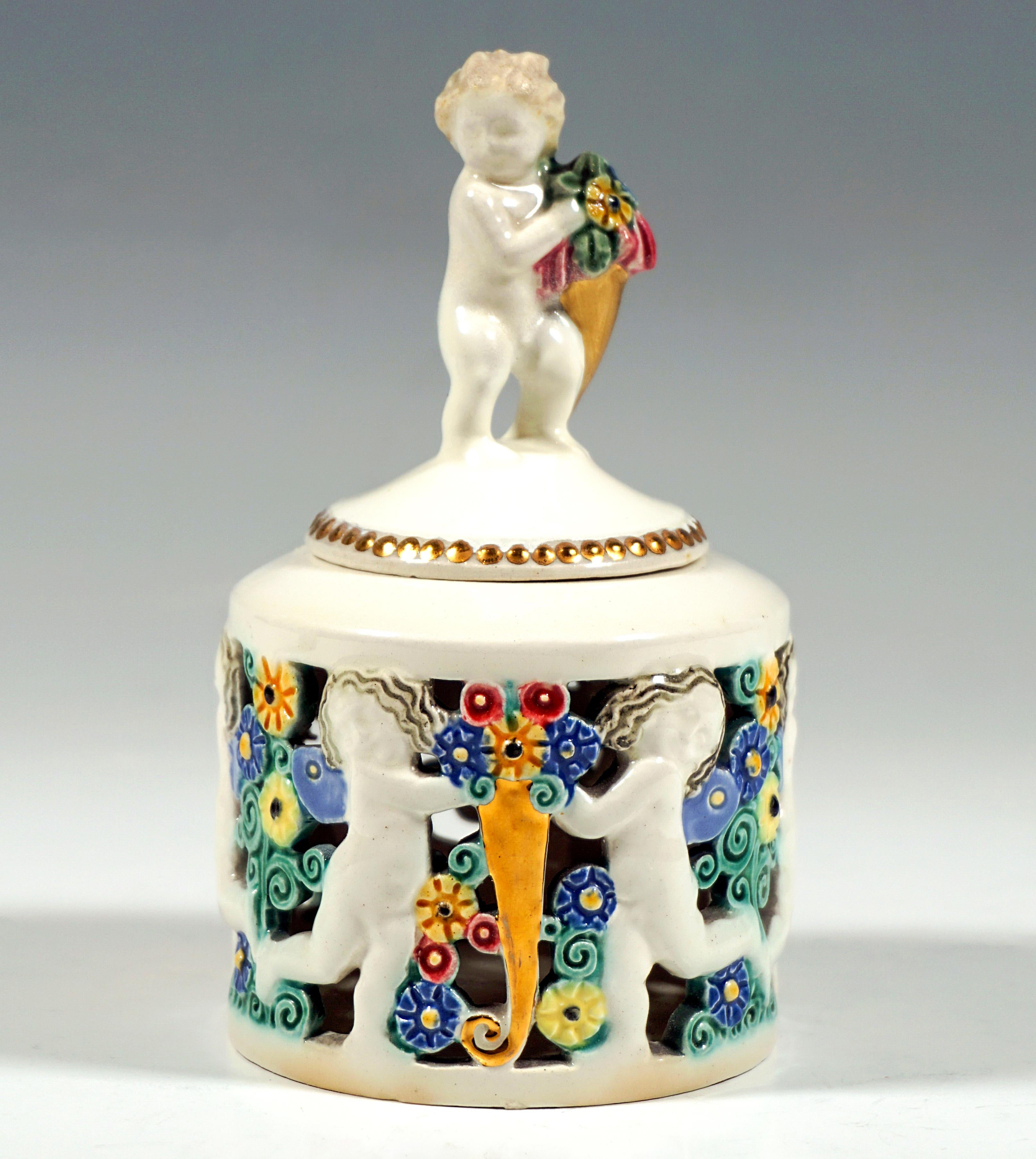Delicate Art Nouveau ceramic piece:
Cylindrical vessel with an openwork relief of running putti, in between colourfully glazed flower bouquets and golden cornucopias with blossoms, the slightly domed, round lid decorated with a golden pearl string