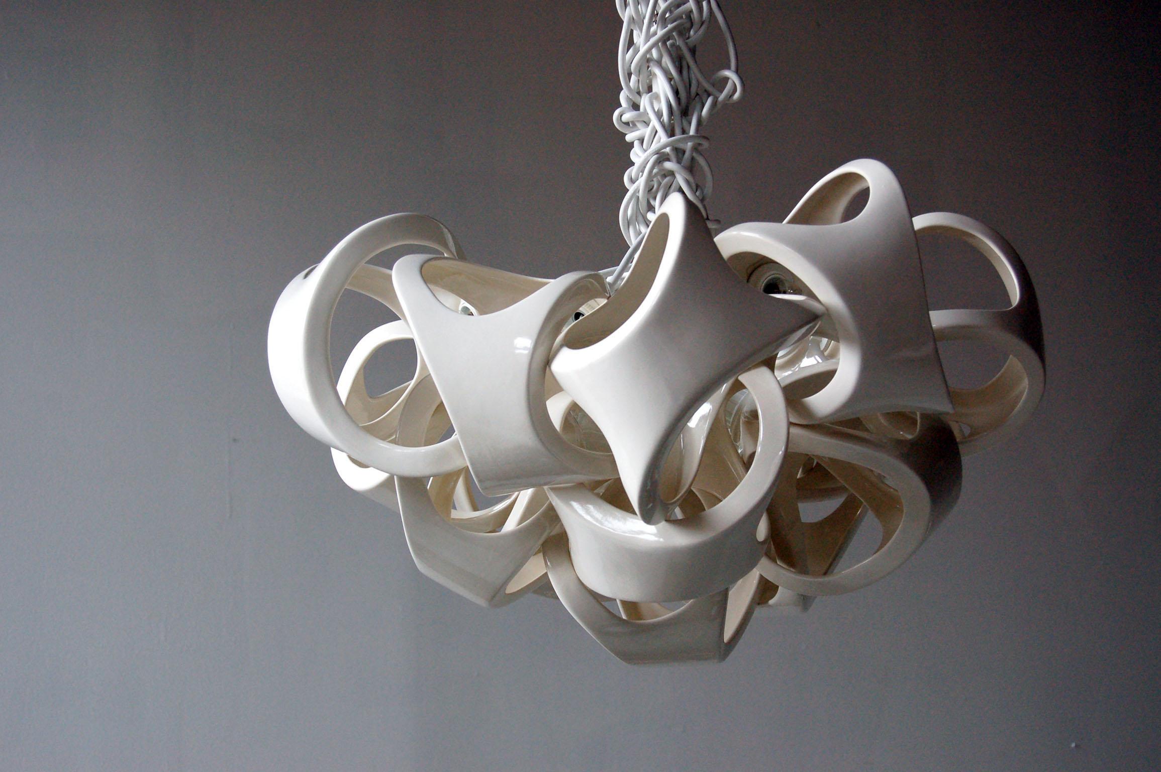 An organic grouping of 15 slip cast ceramic shades. The bone like forms nest together to form a sculptural fixture with dramatic shadows. White knotted cords accentuate the organic nature of the Ceramic lamp series. Overall length of piece can be