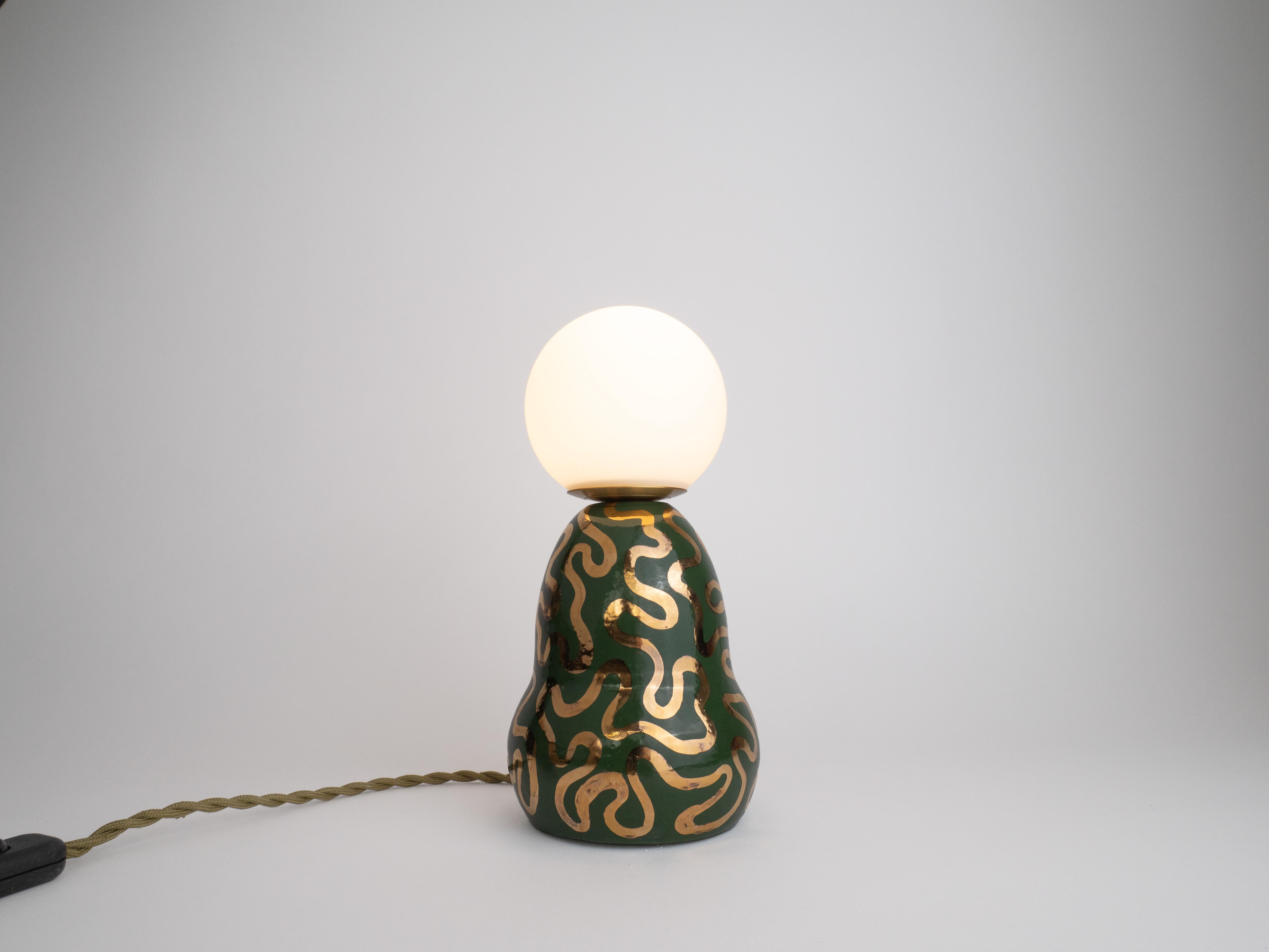 Cette base est recouverte d'une riche glaçure vert foncé, contrastée par un motif ludique de perles dorées qui s'enroule autour d'elle, ajoutant une touche d'élégance et de fantaisie. La combinaison de l'abat-jour en verre doux et translucide et de