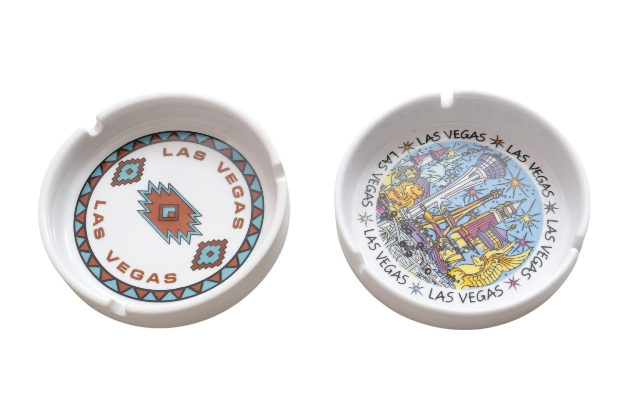 A pair of white ceramic Las Vegas souvenir ashtrays. One is a Hilton ashtray with a southwestern style design in teal and rust circa 1995. The other features various iconic tourist landmarks from around the city. Both have three rounded cigarette