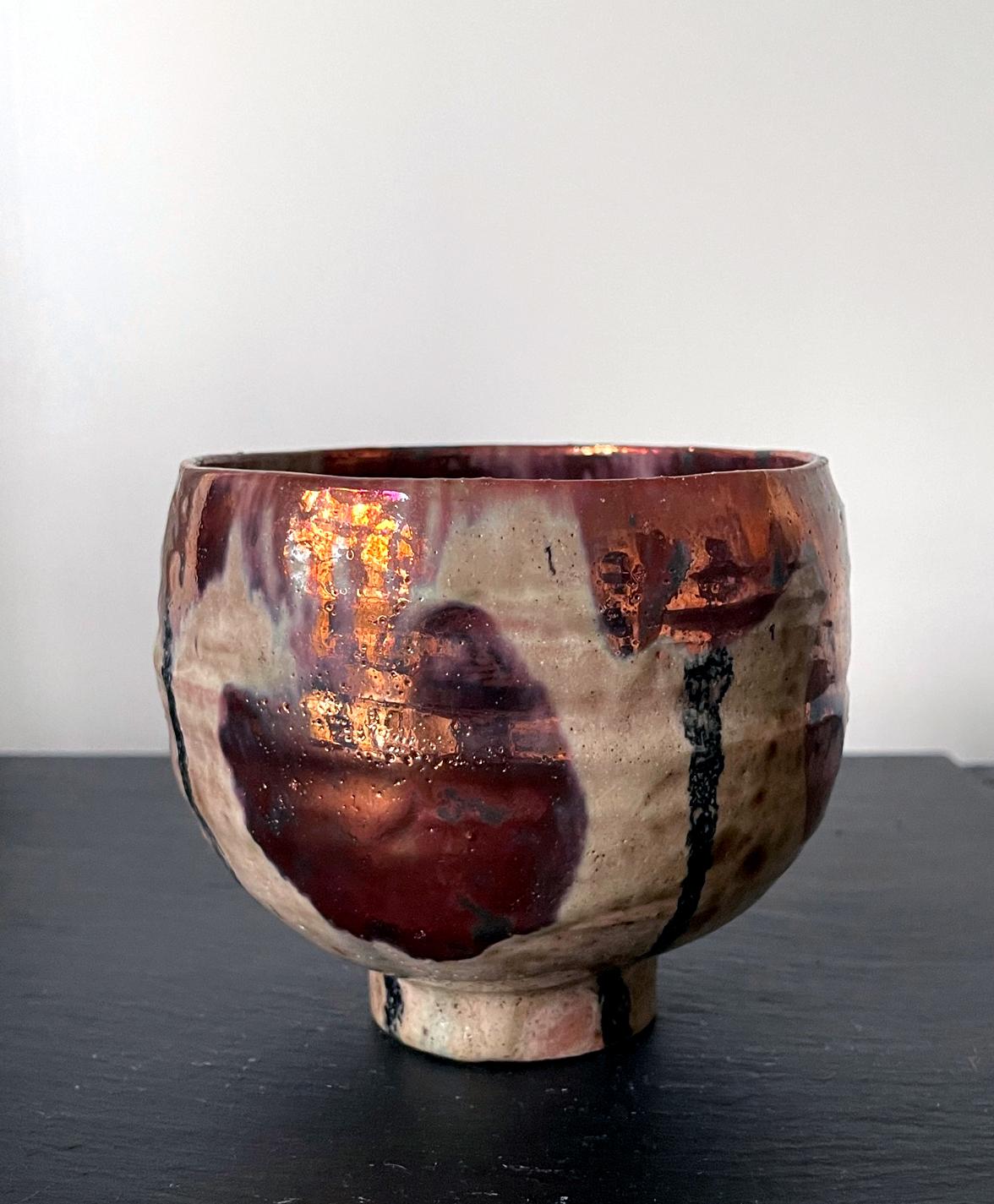 A ceramic lusterware tea bowl by American artist and studio potter Beatrice Wood (1893-1998). The piece, circa 1980s, is of an organic form typical of a Japanese tea bowl. The surface was applied with splashes and drips of a brilliant metallic