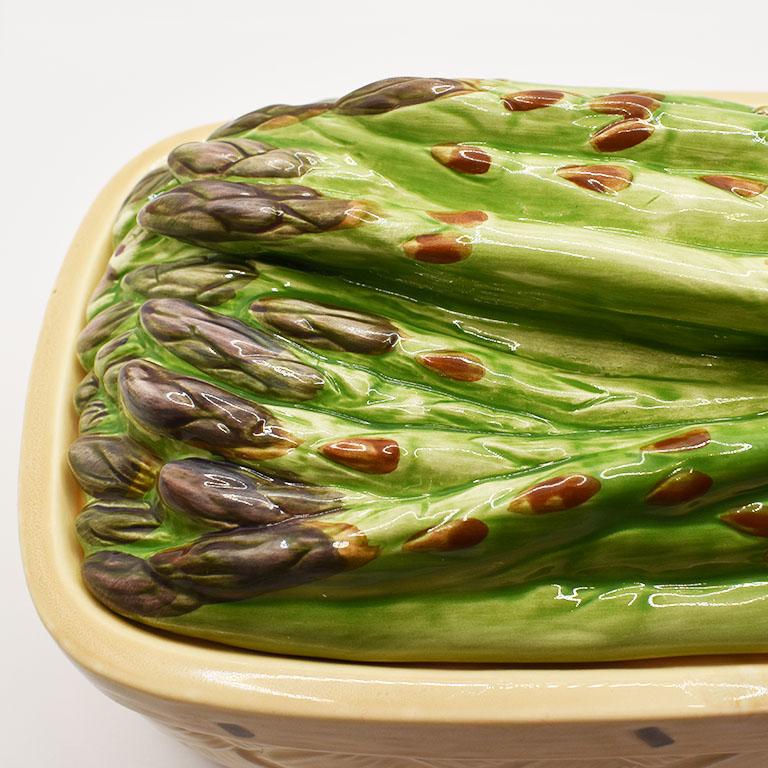 A ceramic majolica serving dish with an asparagus motif lid. This lovely piece will bring a little whimsy to your next dinner party. Created from ceramic, this dish resembles a woven brown woven basket. The lid resembles asparagus sprigs and is