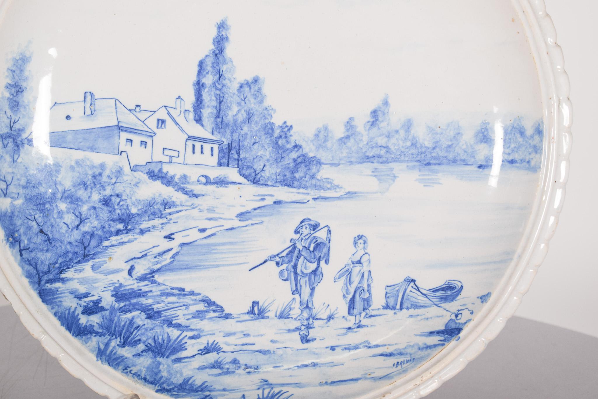 Assiette en céramique, décorée en bleu sur blanc, d'un paysage de campagne et de rivière.
En bas, deux personnages marchent près de l'eau : un homme portant des vêtements traditionnels et un châle sur le dos, et une femme qui le suit de près,