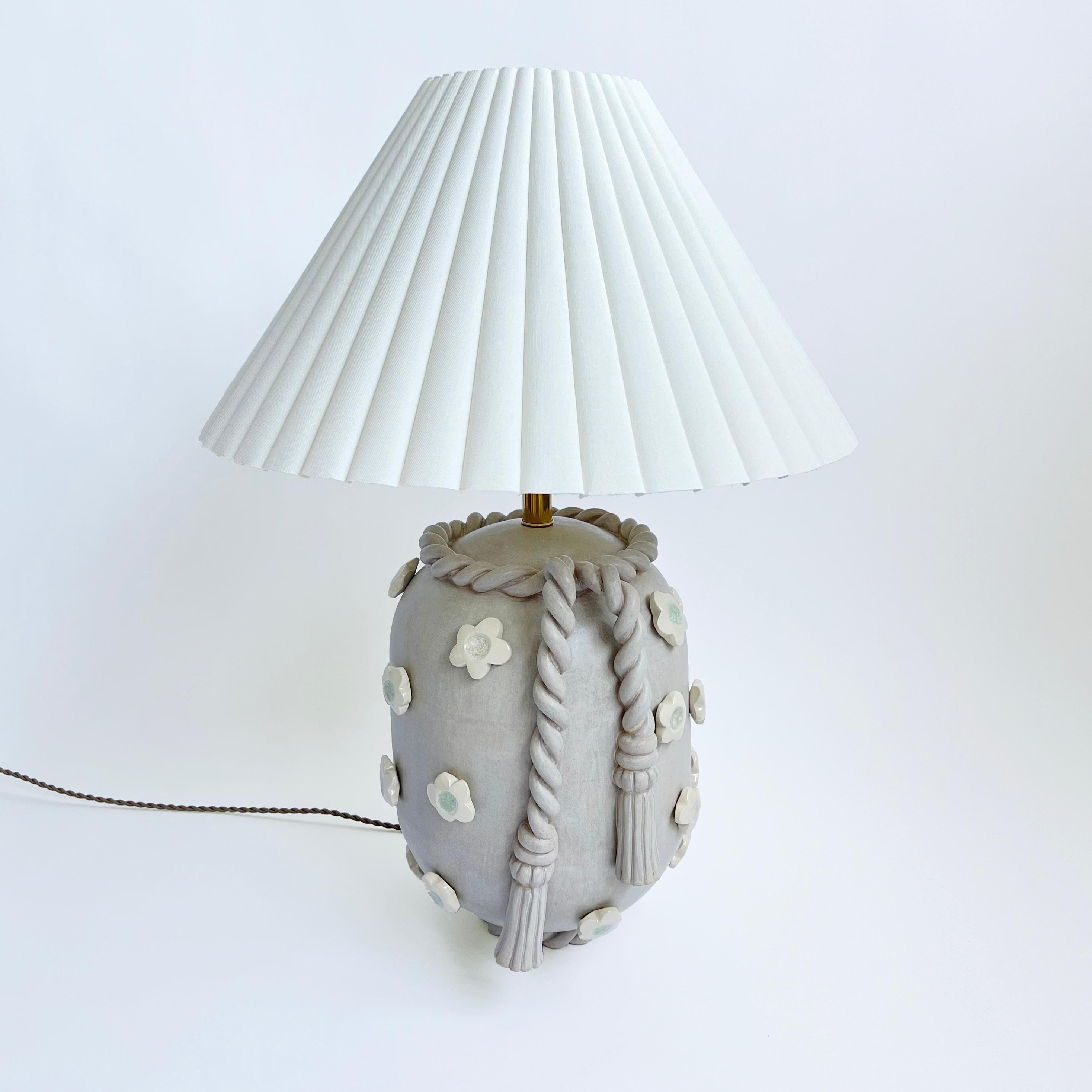 Inspired by architectural details from southern Spain, this ceramic stoneware lamp features hand-sculpted trompe l'oeil tassel detail and hand-carved white stoneware applique flowers inlayed with glass. Finished in light gray satin glaze with brass