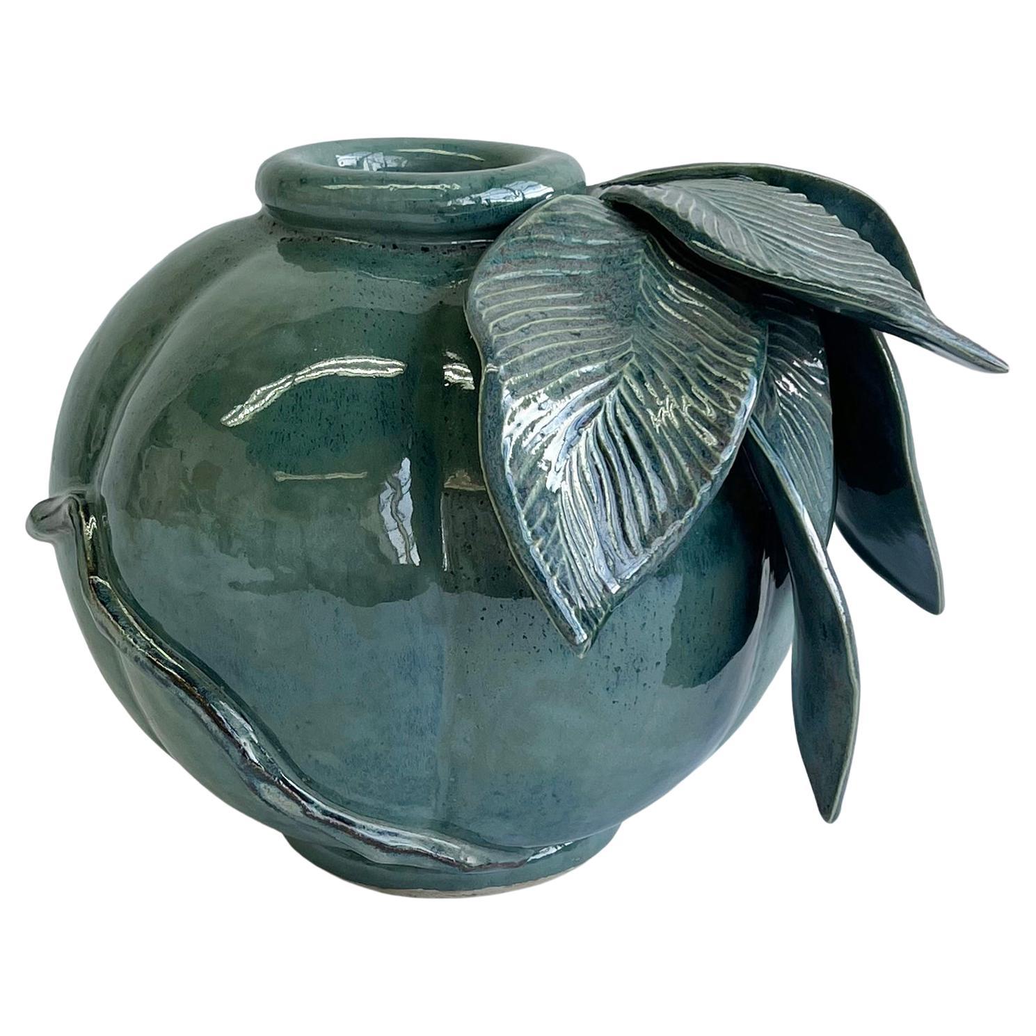 One of a kind, hand sculpted ceramic stoneware vase in glossy, variegated blue glaze with green undertones. This substantial vase is punctuated by hand-carved, oversized, undulating leaves and an angular, wrap around vine. Subtle color variation