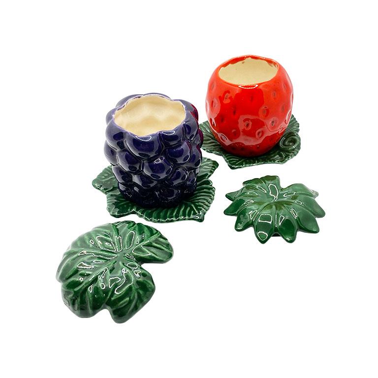 A pair of ceramic jam jars in red and purple. This set will be a fun way to serve breakfast. Each jar sits upon a green cabbage leaf plate. One jam jar depicts a grouping of grapes, while the other depicts a strawberry. Each piece has a leafy lid