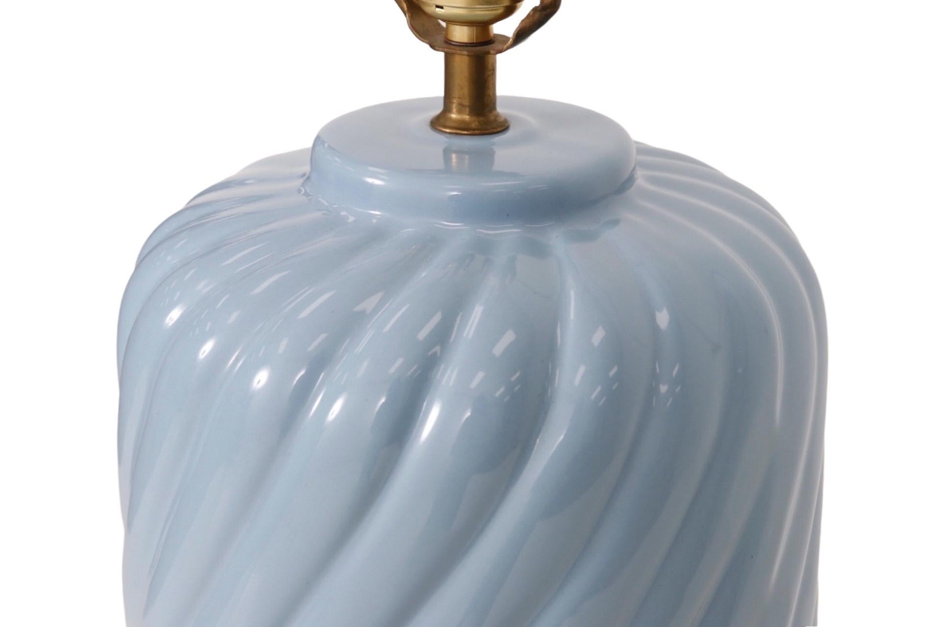 A cylindrical ceramic table lamp in periwinkle blue. Bevelled lines swirl diagonally around the body of the lamp. The central column and harp holder are brass.
  