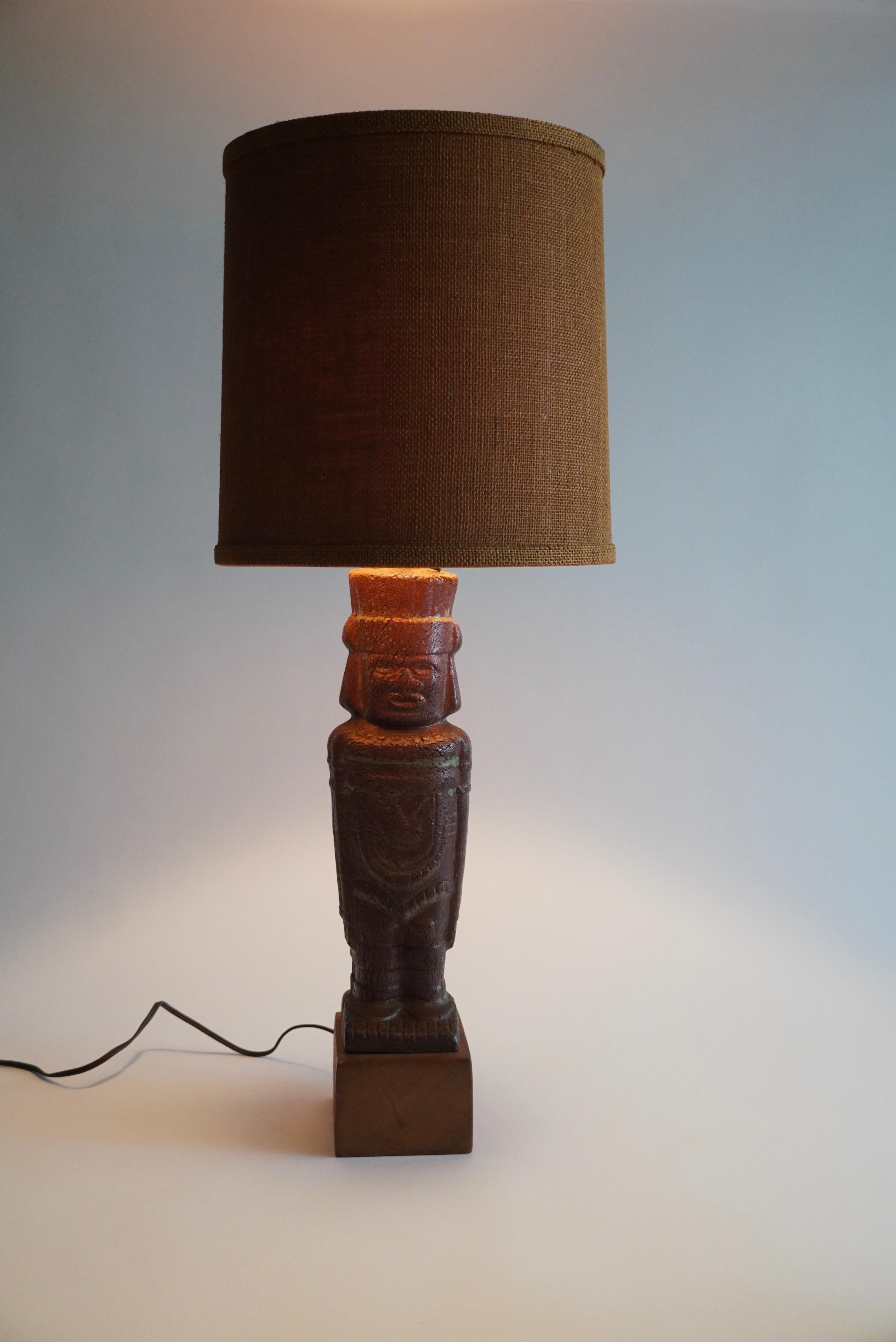 1950s Ceramic table lamp with a stylized Aztec figure with its original burlap covered lamp shade. The figure is done in a rich earth toned brown crackle glaze with some green accents in the glaze and is mounted a top a wooden base.