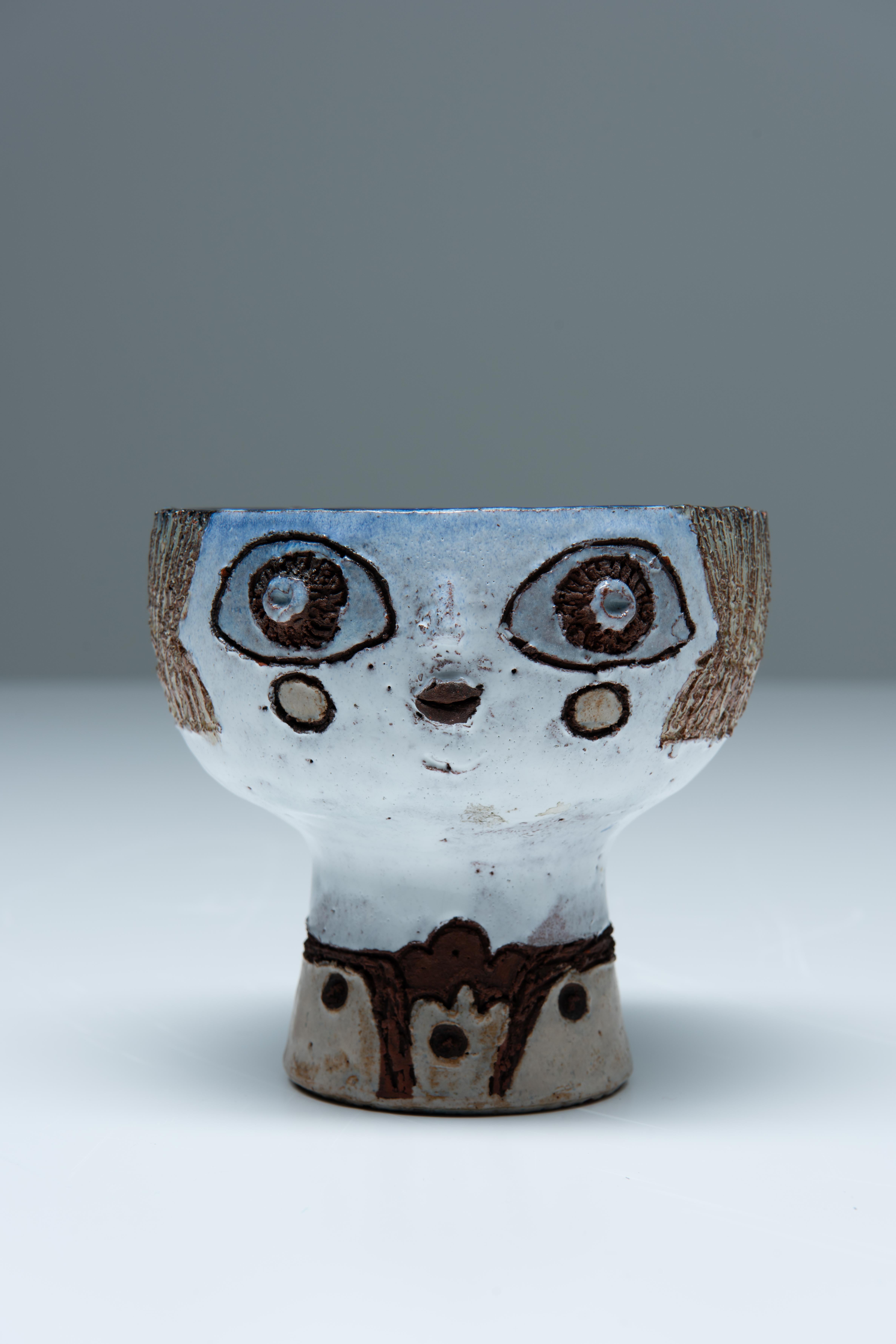 Short pedestal vase with human-like face on front by Belgian ceramist Elisabeth Vandeweghe from the 1970's.
Vandeweghe is still active in the art world. Her work is very figurative and has a deep nice colored glazing. 
Together with other ceramists,
