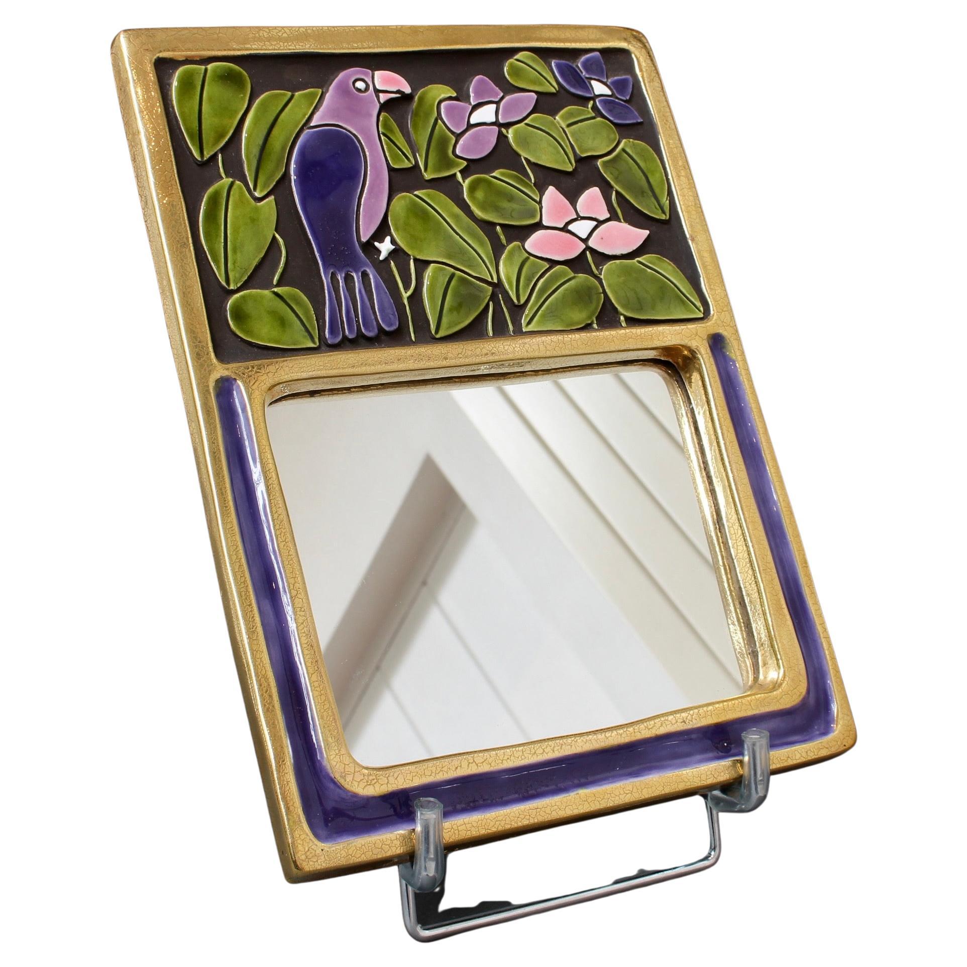 Ceramic wall mirror with purple and green glaze and stylised bird (circa 1970s) by Mithé Espelt. A delightful whimsical wall mirror decorated with a parrot perched amongst the flowers and greenery situated above the rectangular mirror. The mirror is