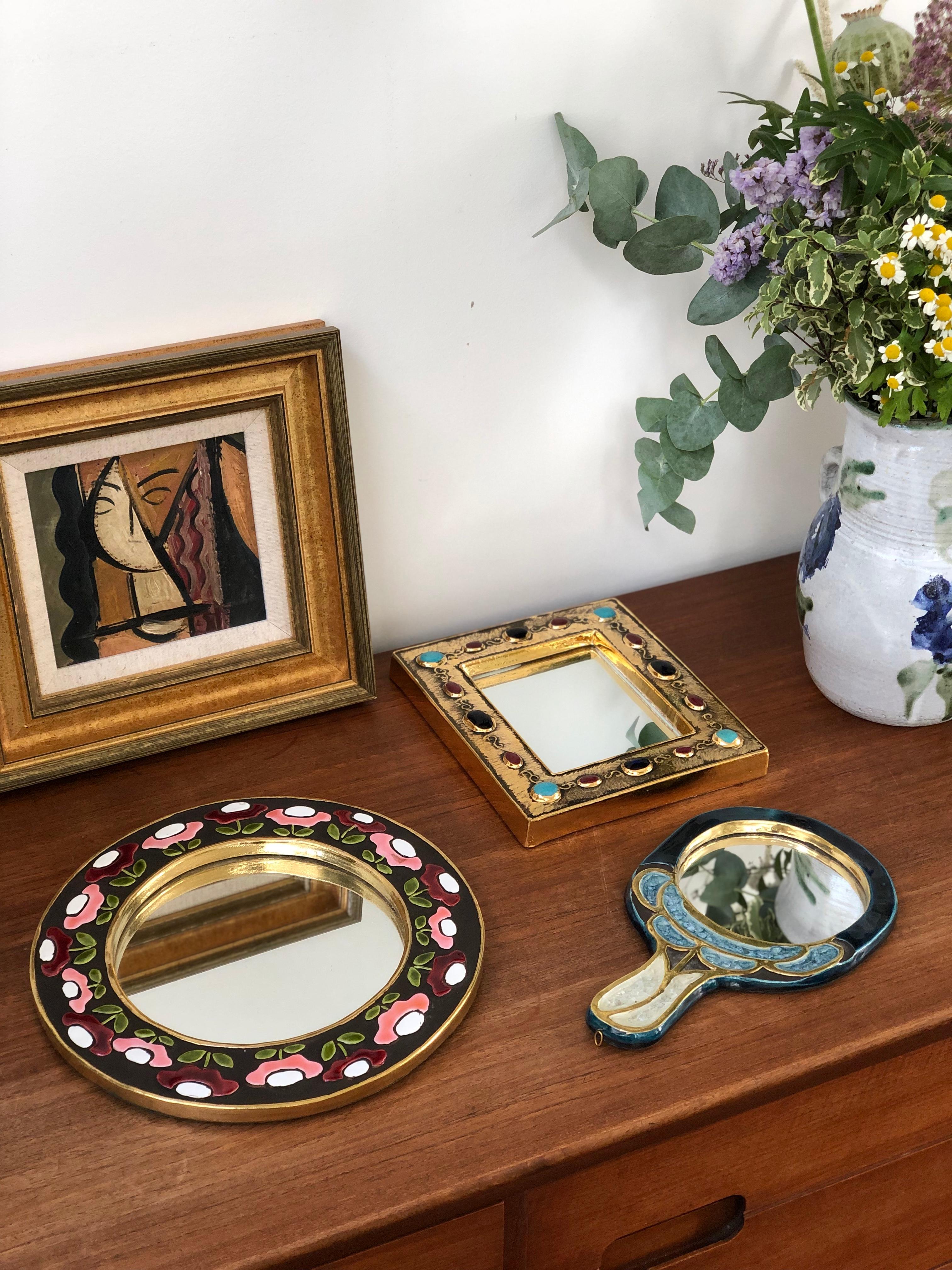 Ceramic wall mirror with flower motif (circa 1960s), by Mithé Espelt. A porthole-shaped decorative wall mirror delights the eye. The inner and outer frames are in a stunning gold craquelure. The enamelled flowers are superimposed over a chocolate