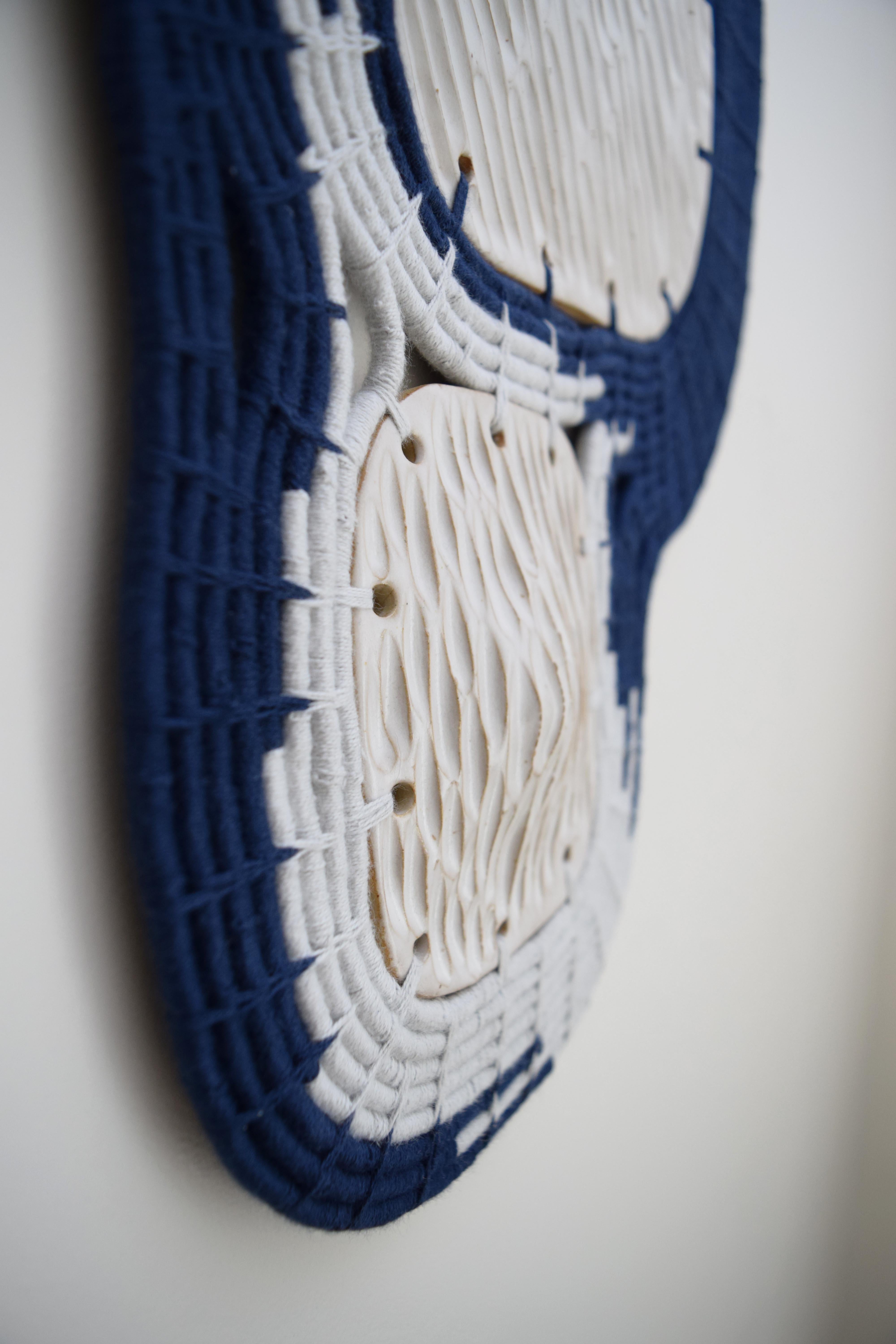 Paneles de cerámica esmaltada blanca con textura rodeados de algodón tejido en azul marino y azul pálido. Los paneles tienen dimensión, creando una superficie ondulada. El cable para colgar se incluye en la parte posterior. 

18