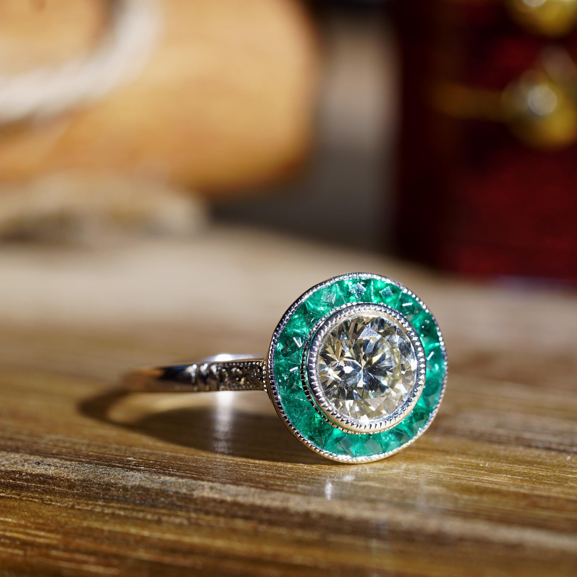 An Art Deco inspired target ring, boasting a central bezel set GIA certified 1 carat diamond with a color of N and clarity of VVS1. The central diamond is surrounded by a halo of channel set French cut emeralds. The round diamonds on shoulders add