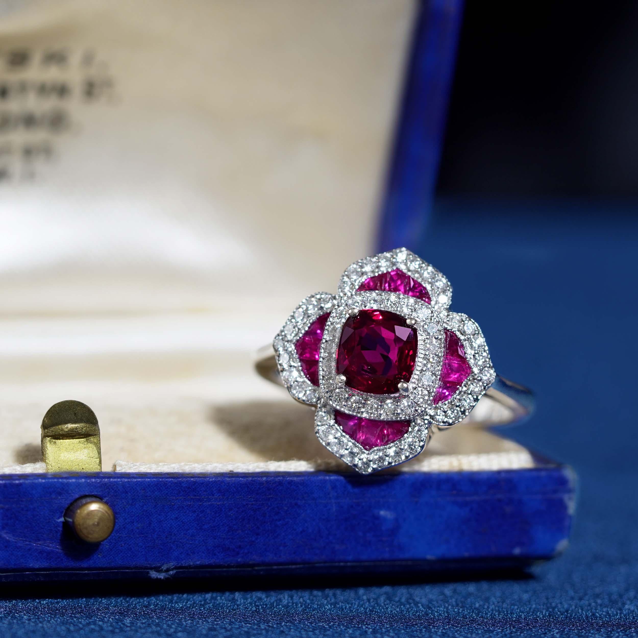Süß und raffiniert. Dieser fein gearbeitete Ring aus 18 Karat Weißgold enthält einen kissenförmigen Rubin von 1,02 Karat (ca.) mit Akzenten aus runden Diamanten im Brillantschliff und Rubinblüten im französischen Schliff. Dieser atemberaubende Ring