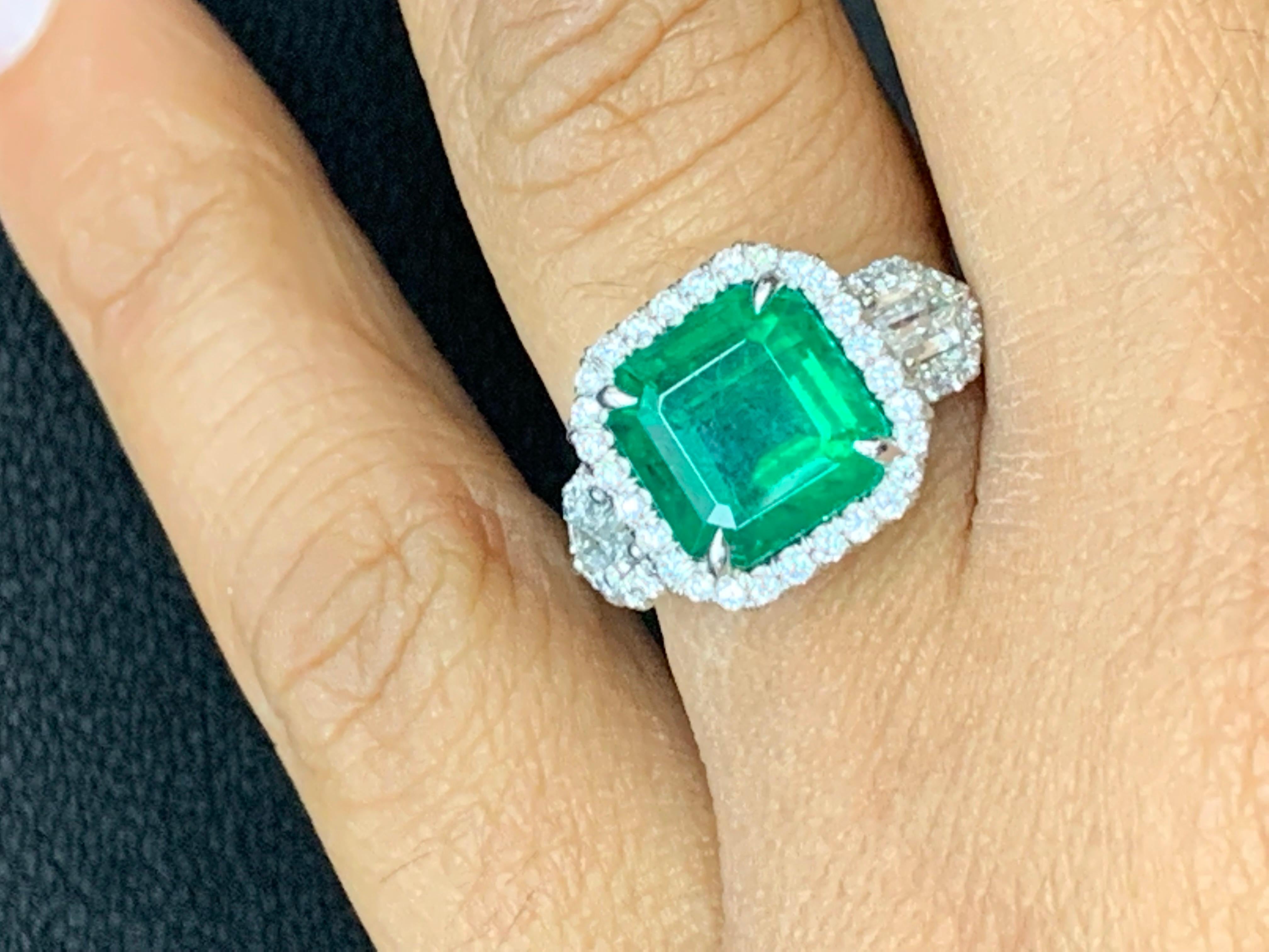 An antique style engagement ring showcasing a 3.69-carat emerald cut emerald, surrounded by a row of brilliant cut diamonds and two brilliant bullet diamonds on either side. The piece is finished with a brilliant diamond halo and is set in an