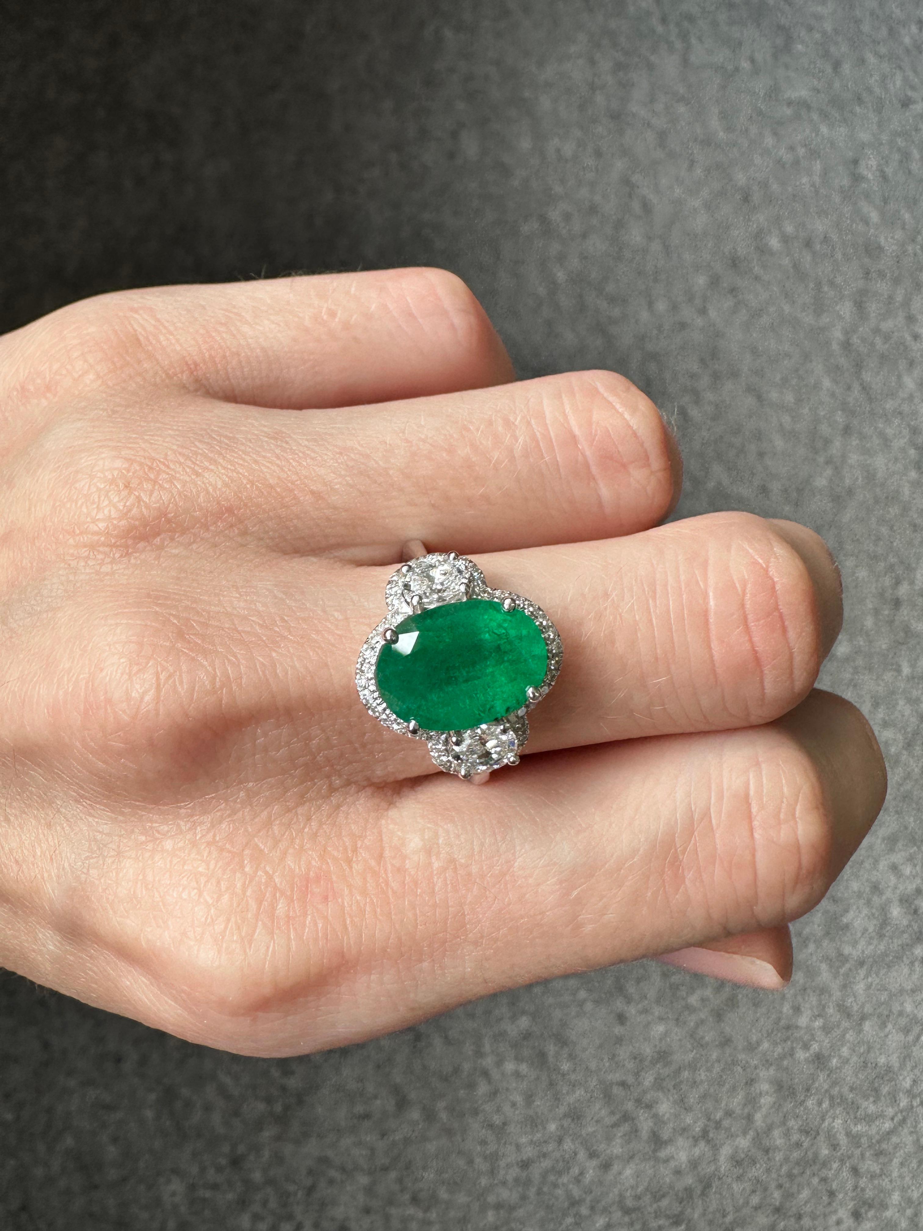 A classic 4.06 carat Zambian Emerald three stone engagement ring, with 0.61 carat oval shaped Diamond side stones and a Diamond halo. The center stone is an ideal vivid green color, with great luster and transparency. The stones are set in solid 18K