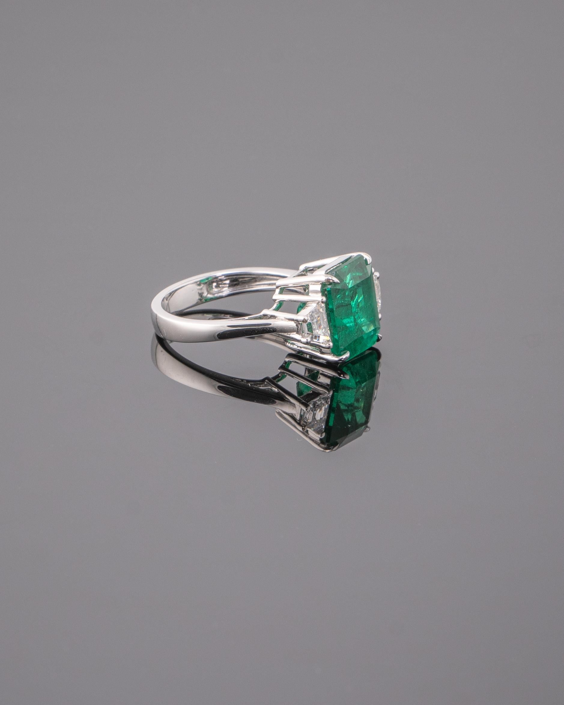 A stunning three stone engagement ring, with a 6.43 carat transparent natural Zambian Emerald centre stone and 2 half-moon side stone diamonds. The emerald is of great lustre and has an ideal colour. All set in 18K white gold. Currently a ring size