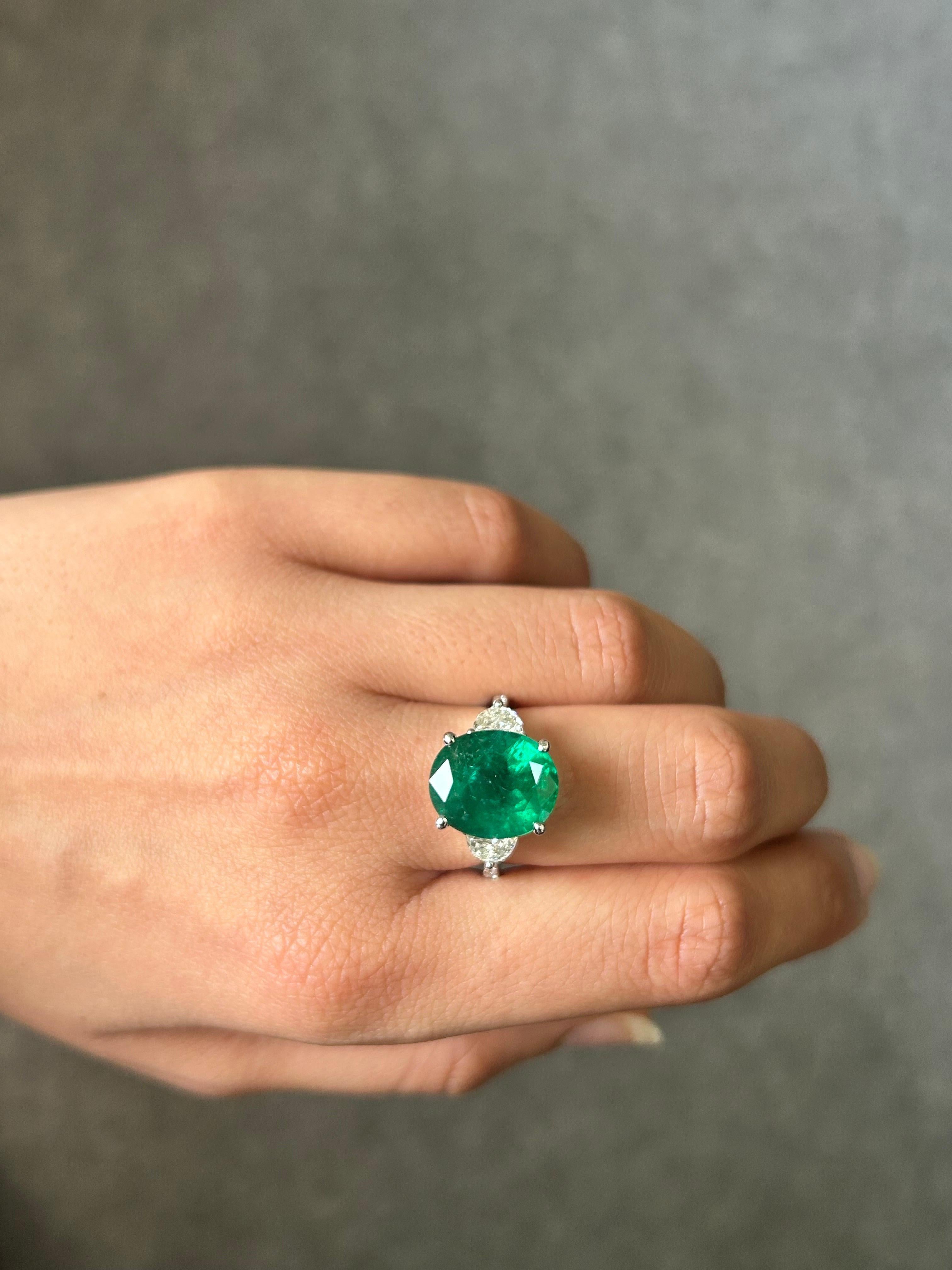 A beautiful 7.12 carat oval shaped natural Zambian Emerald three-stone engagement ring, with 0.62 carat half-moon diamonds and 0.13 carat round diamonds on the band. The center stone is of top quality, with a stunning vivid green color and is