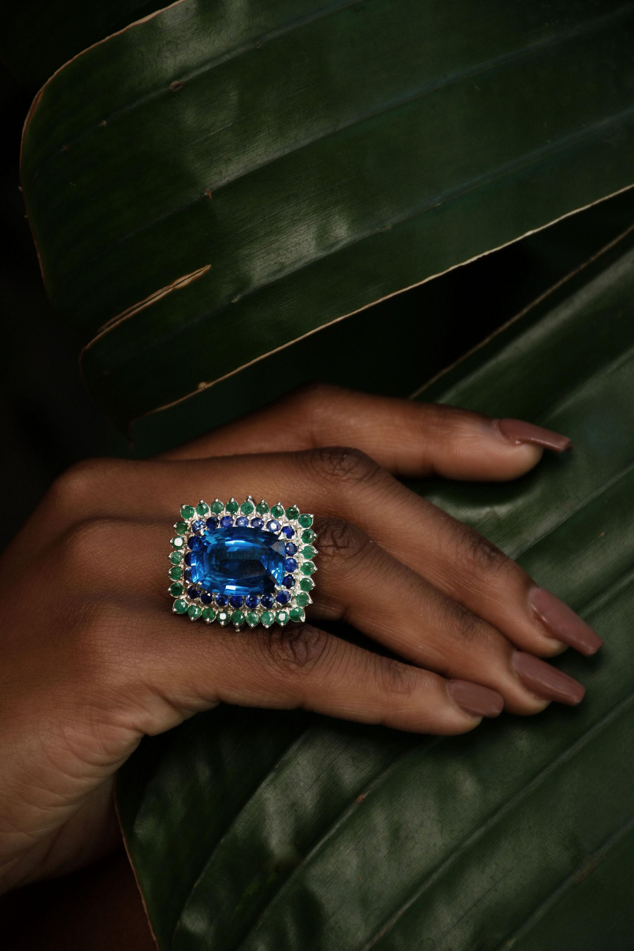 Custom Platinum Blue Topaz Cocktail Ring with Emerald and Sapphire Halo

This custom-designed platinum cocktail ring is a statement of unparalleled elegance and bold sophistication. At its center, a 13-carat, 18.2 x 13.2mm cushion-cut natural blue