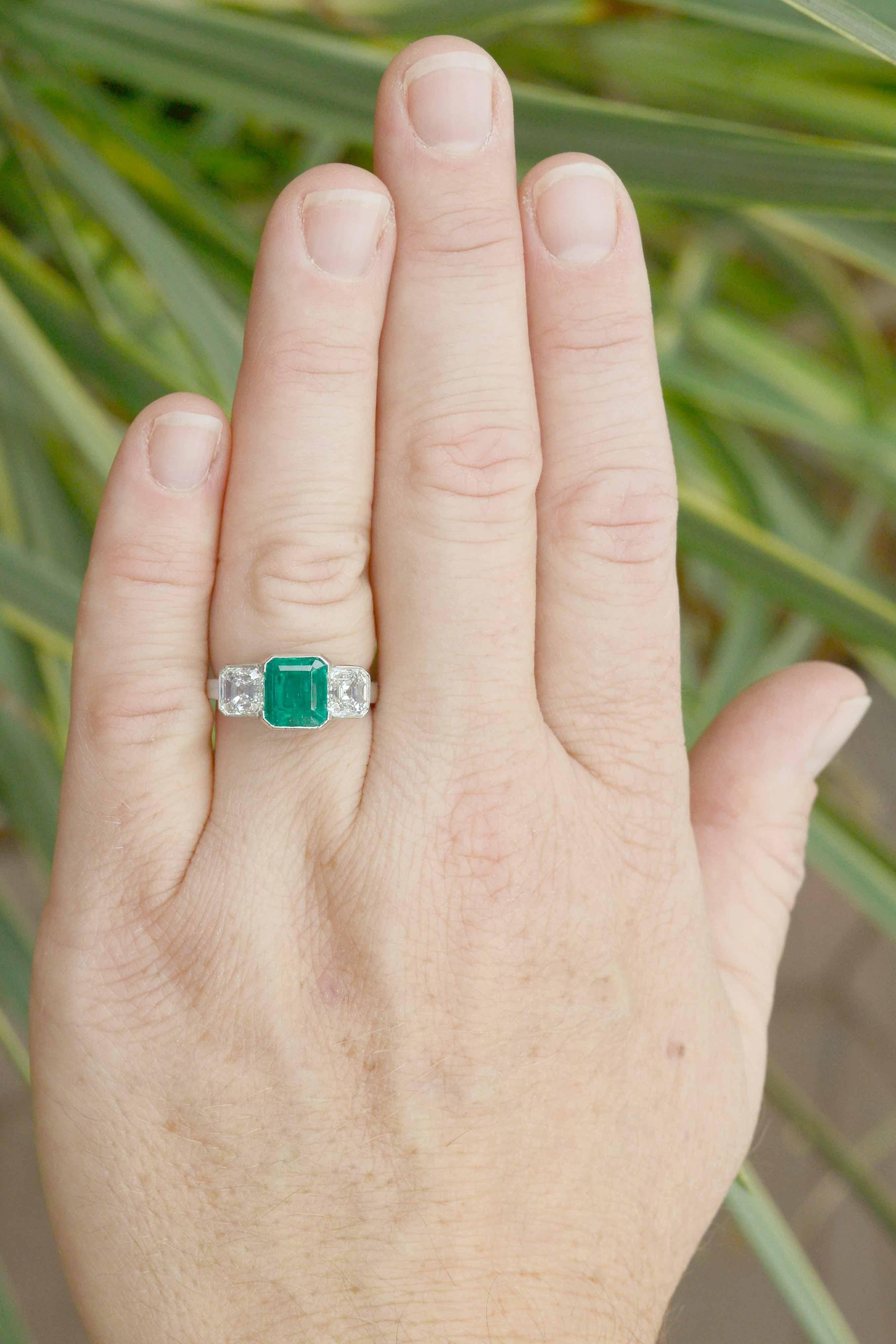 The pedigree of this Colombian emerald 3 stone Art Deco engagement ring is impressive. The sleek design starring a natural emerald cut Muzo mine emerald of 2 1/2 carats of an intensely saturated green that glows from within, having excellent
