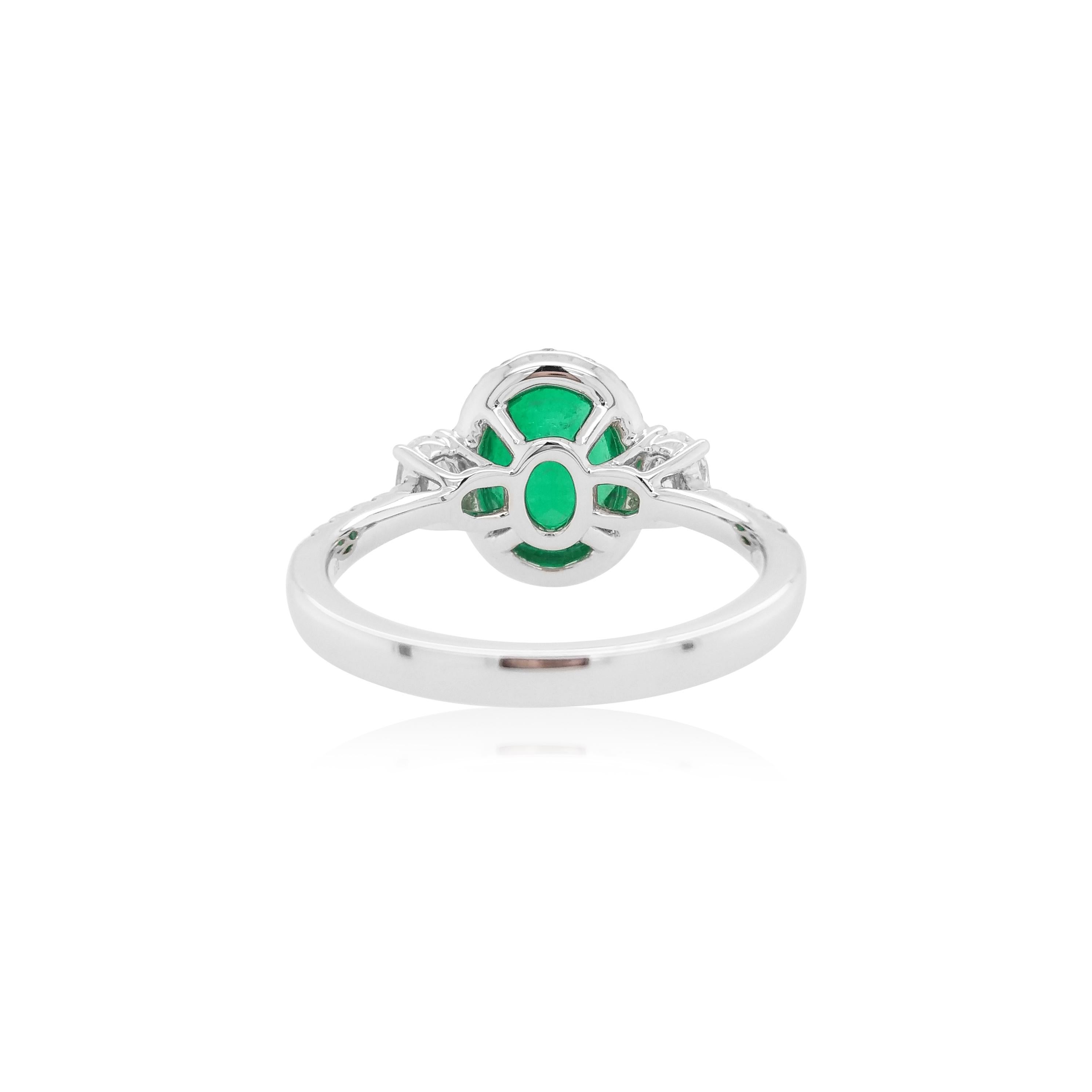 This stunning ring features a vibrant oval-shaped Colombian Emerald at its forefront, accentuated by a delicate white diamond halo surrounding it, and a pair of oval-shaped scintillating white diamonds on the shoulders. Set in 18 Karat white gold to