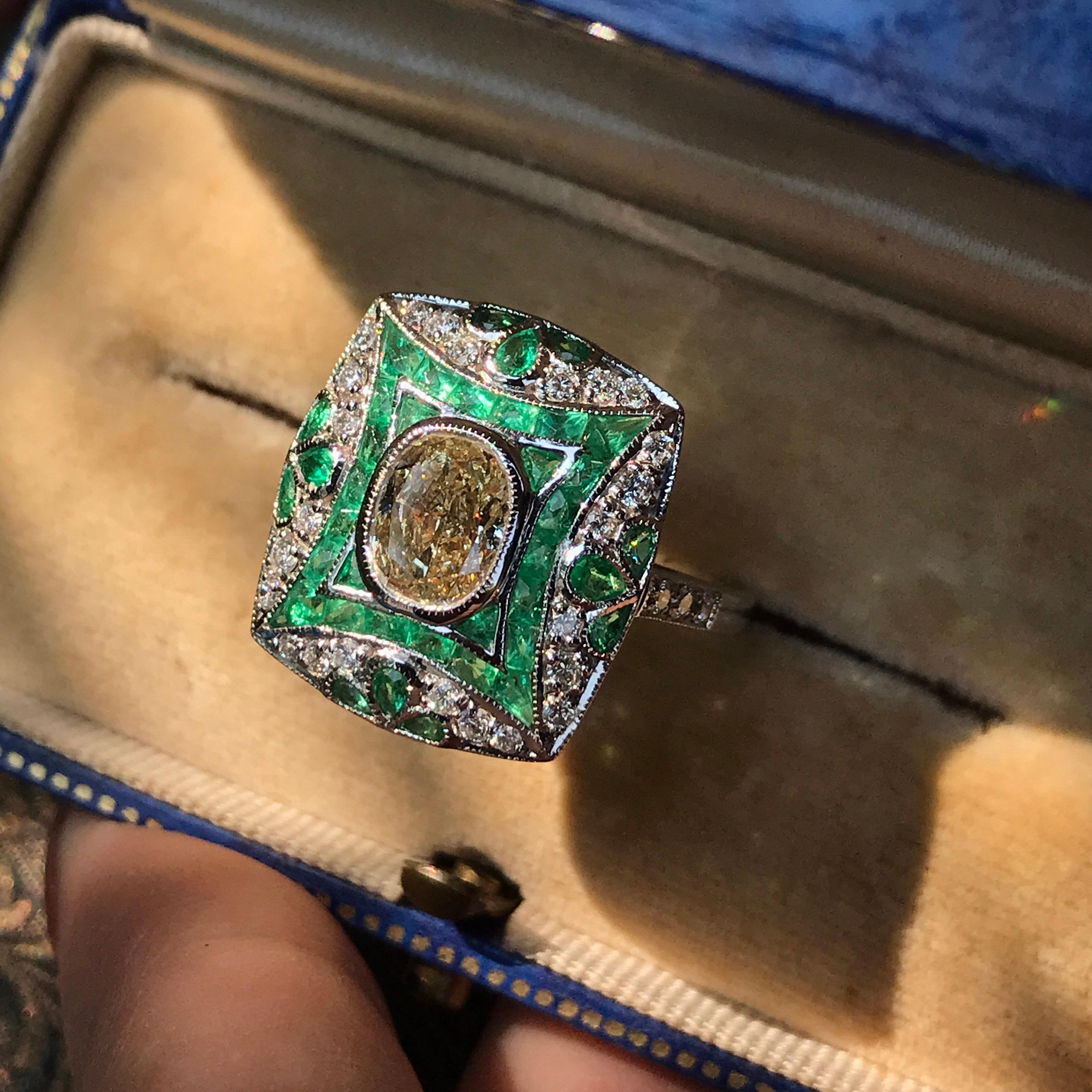 In this artful Vintage inspired ring, an oval shaped diamond glitters from the center of 18k white gold setting accented by French cut emeralds and round diamonds. The emeralds were add for its lively green leaves motif, producing a beautiful and