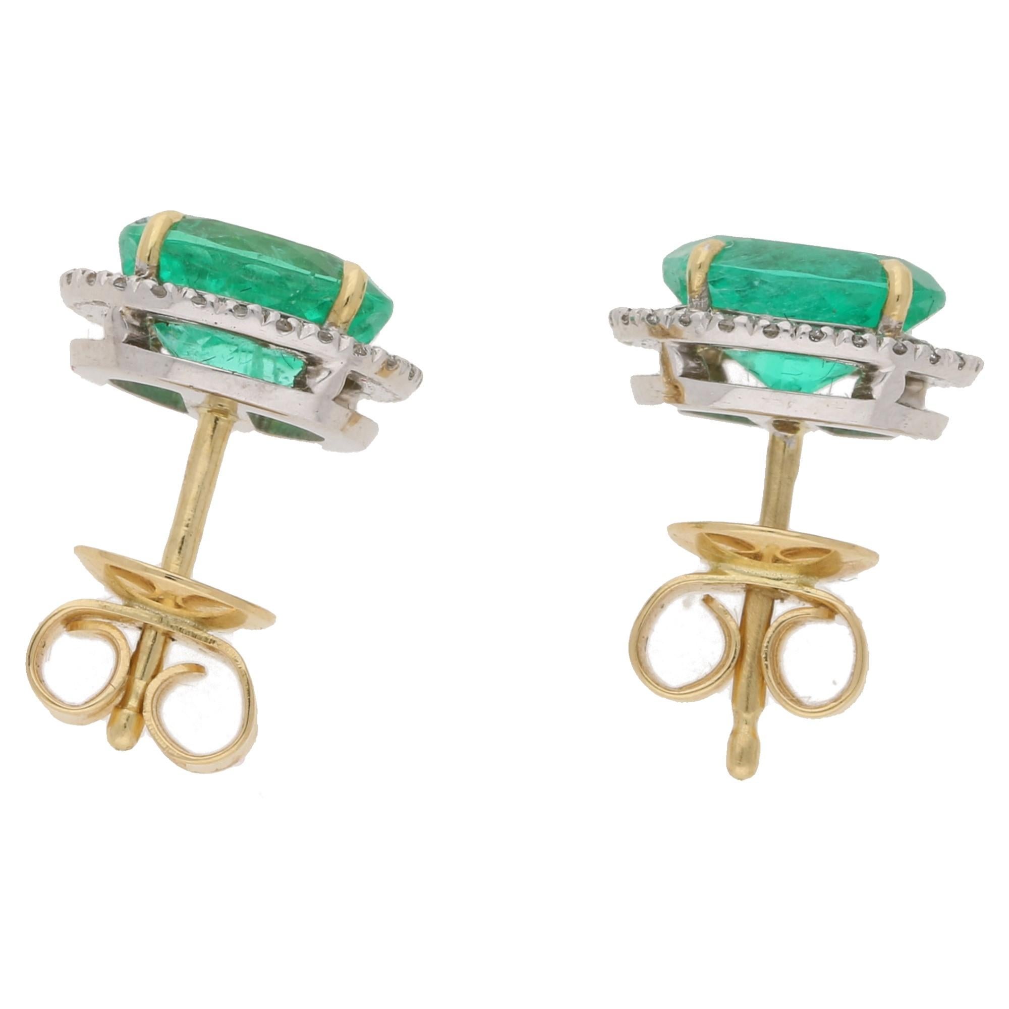 A stunning pair of emerald halo stud earrings set in 18ct gold featuring 1.36ct and 1.28ct oval cut Columbian emeralds independently certified by GCS, set in four claw yellow gold settings with a diamond halo surround set in white gold to post and